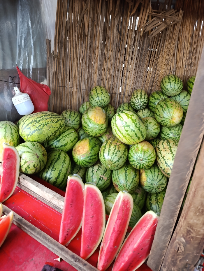 The image shows a vendor selling watermelons in Githurai, Kenya, located at coordinates -1.203 latitude and 36.918 longitude. A large pile of watermelons is displayed on a wooden stand, positioned under a thatched roof overhang. The stand is situated against a wooden wall, and the watermelons are arranged haphazardly. Several watermelons are cut open, revealing their red flesh, and are displayed for sale. There are no people visible in the image. The environment appears to be a bustling marketplace, with the vendor's stand likely part of a larger outdoor market area.