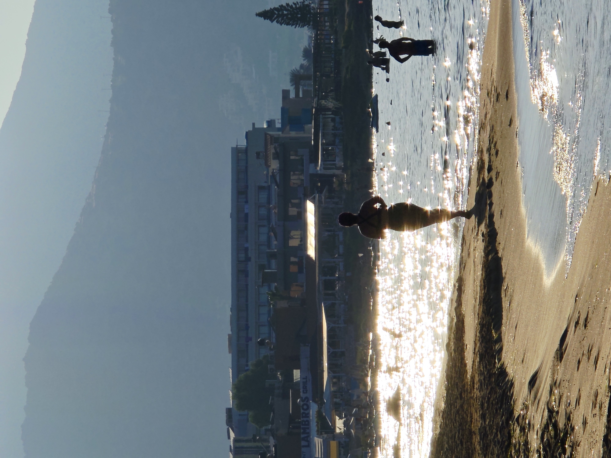 This image depicts a beach scene in the Malia Municipal Unit, Greece. In the foreground, a sandy beach slopes down to the water, with gentle waves lapping the shore. The water is sparkling with sunlight, creating a shimmering effect. Several people are visible in the water, some swimming and others standing. In the mid-ground, a woman in a swimsuit is walking along the shoreline, her back to the camera. Behind the beach, a row of buildings lines the coast, including what appears to be a hotel and several restaurants with outdoor seating. In the background, a large, hazy mountain range dominates the horizon under a pale sky. A lone, tall evergreen tree stands to the right of the buildings. The overall impression is of a bright, sunny day at a popular tourist destination.