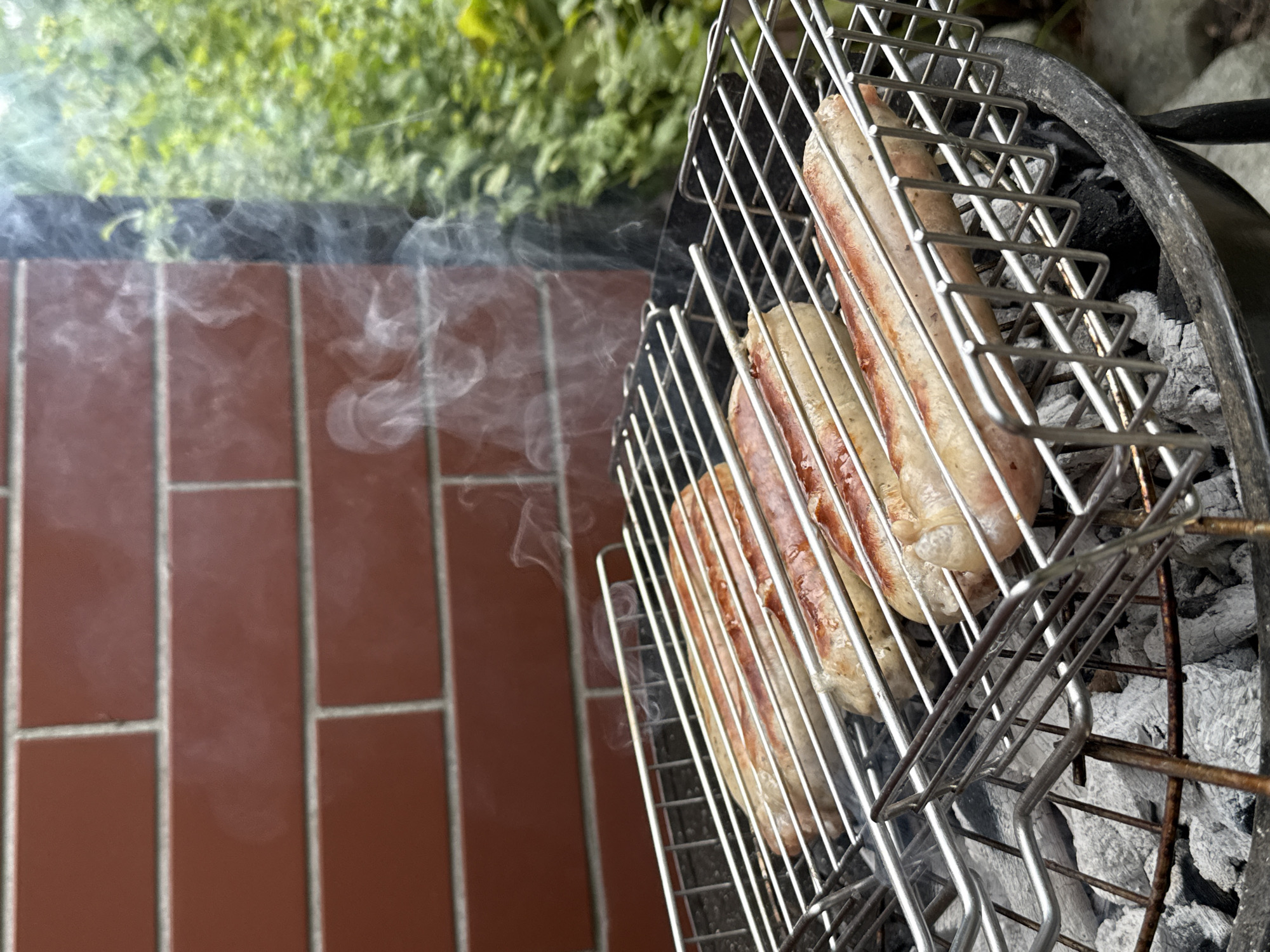 A close-up shot captures four sausages sizzling on a metal grill over hot coals. The grill is positioned outdoors, with a brick wall in the background and lush green foliage to the right. Wisps of smoke rise from the sausages, indicating they are actively cooking. The scene appears to be a casual outdoor barbecue, likely in a residential setting within the city of Wrocław, Poland. The lighting suggests it is daytime, and the overall atmosphere is one of a relaxed, home-style meal preparation.
