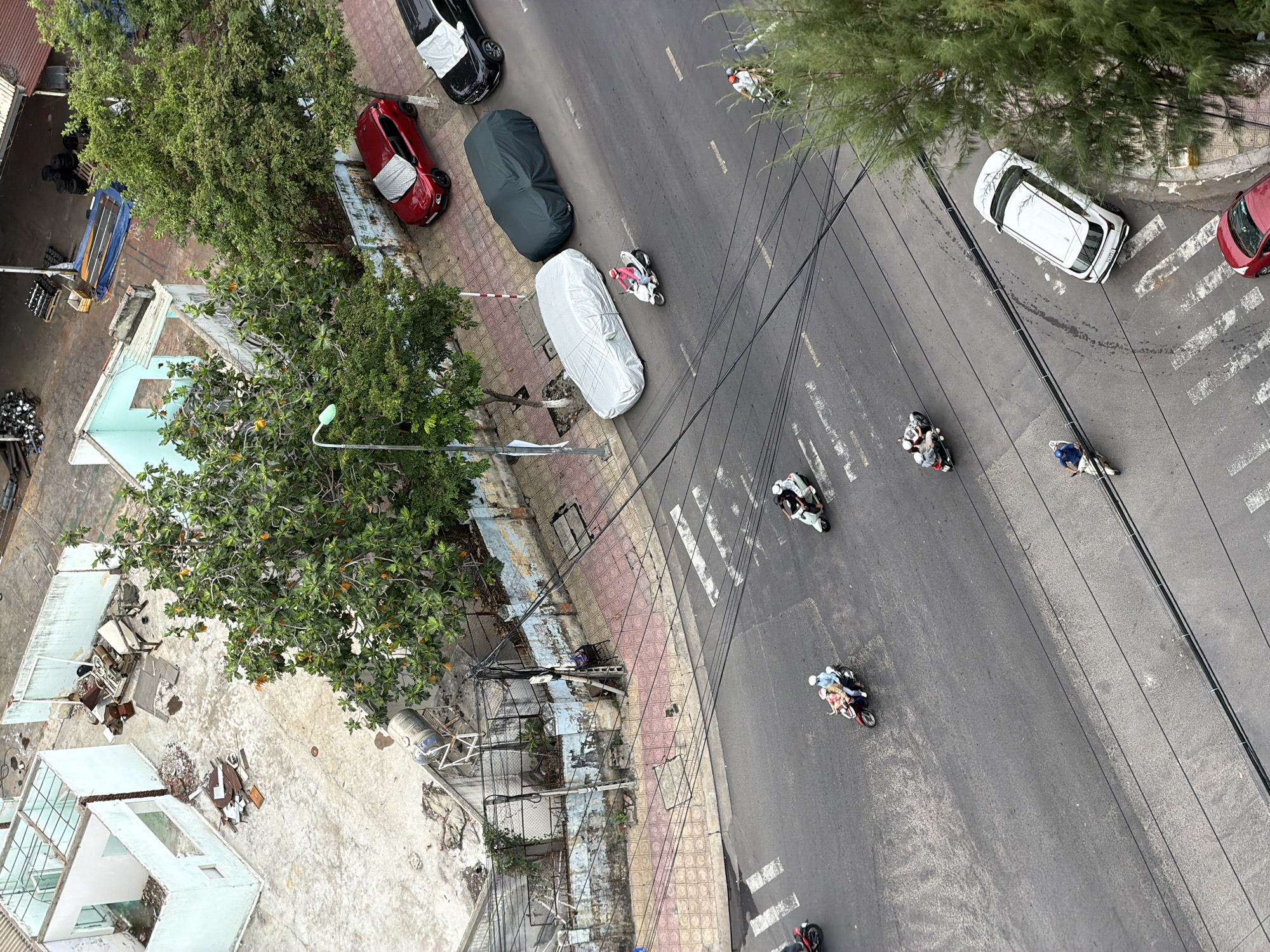 This aerial view captures a street scene in Nha Trang, Vietnam. The image shows a two-lane road with several vehicles and pedestrians. On the right side of the road, there is a red car, a black car covered with a tarp, and a light grey car also covered with a tarp. Further down the street, a white SUV is parked. Several motorbikes are visible, with riders, some carrying passengers. One motorbike has two people on it, another has a single rider, and a third has two people including a child. On the left side of the road, there is a sidewalk with a street lamp and trees. Behind the sidewalk, there are buildings, some of which appear to be under construction or in a state of disrepair, with exposed walls and debris. The road surface is asphalt, and there are white pedestrian crossing lines painted on it. Overhead, a complex network of electrical wires is suspended. The general environment appears to be an urban setting with mixed development.