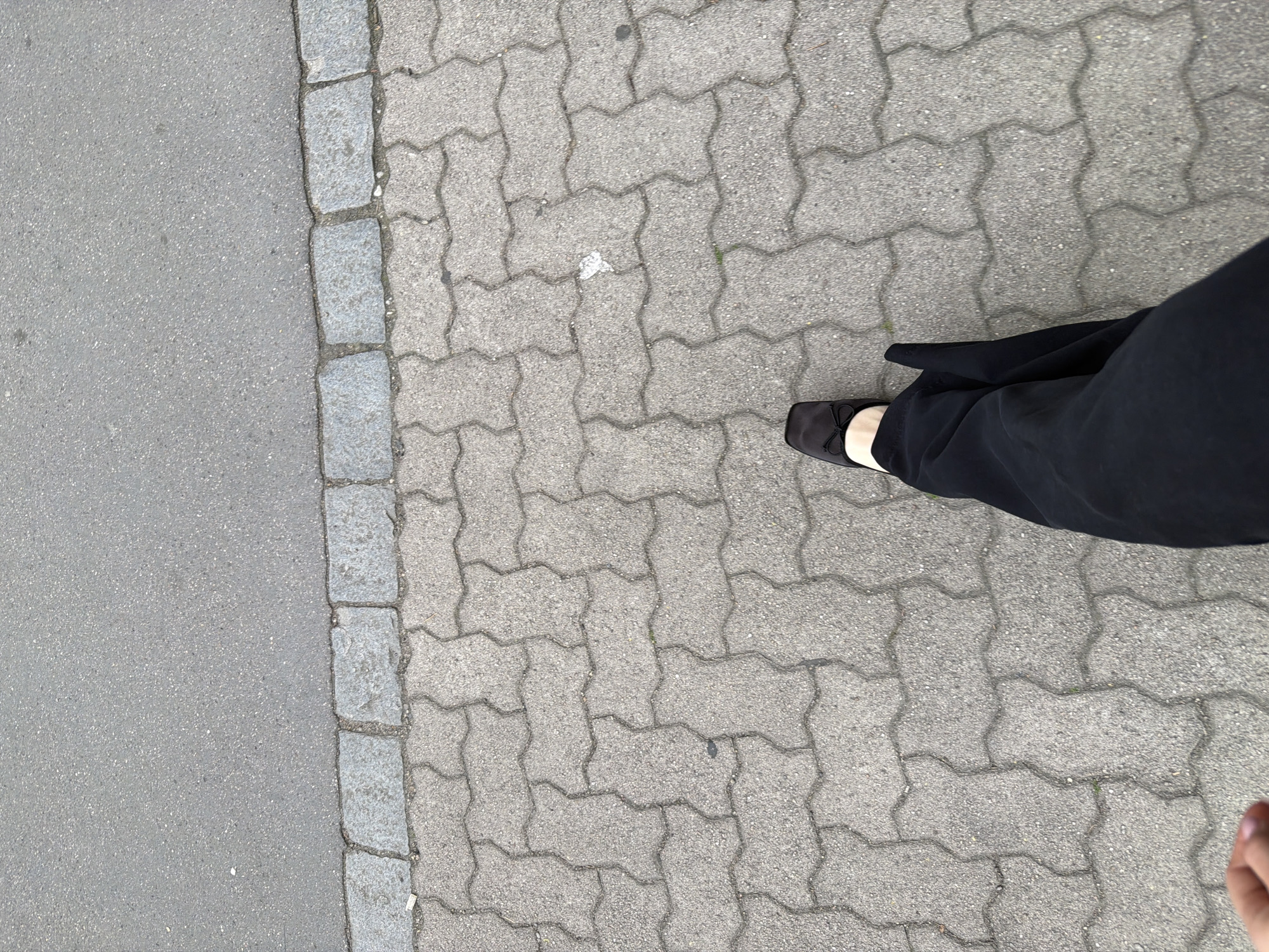 The image shows a person's legs and feet from a first-person perspective, walking on a paved sidewalk. The sidewalk is made of interlocking concrete pavers, with a border of larger, rough-hewn stones separating it from an asphalt road. The person is wearing black trousers and black flat shoes with a small bow on the toe. The lighting suggests it is daytime, possibly overcast. The background is a plain asphalt road. There are no other people or significant objects visible in the frame. The location is a city street in Strasbourg, France.