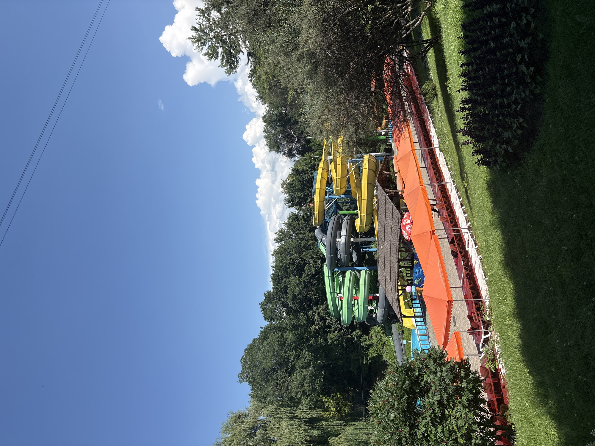 The image depicts a water park on a clear, sunny day. In the background, a multi-level water slide complex is visible, featuring brightly colored tubes in green, yellow, and black. These slides are surrounded by lush green trees. In the foreground, a row of orange sunshades covers lounge chairs and seating areas, suggesting a recreational space. The ground is covered in grass, with some dark, leafy plants visible. A swimming pool is partially visible near the slides. The scene appears to be an outdoor leisure facility. The location is identified as being in Almaty, Kazakhstan.