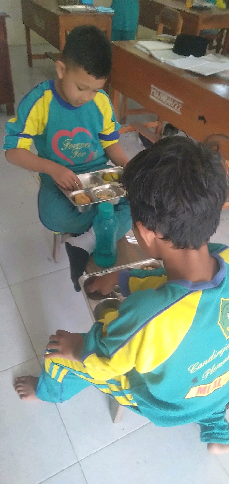 Two young boys, appearing to be of Indonesian ethnicity, are seated on the floor in what seems to be a classroom or dining area in Plemahan, Indonesia. Both boys are wearing matching teal and yellow athletic wear. The boy on the left is holding a metal lunch tray with various food items, including what looks like fried food, rice, and a piece of fruit. He is looking down at the tray. The boy on the right is also looking at a similar lunch tray, and his hand is reaching towards it. A blue water bottle is placed on the floor near the boy on the left. The floor is tiled and appears to be light-colored. In the background, wooden desks and chairs are visible, suggesting an educational setting. The overall impression is of two children having a meal break, possibly at school.