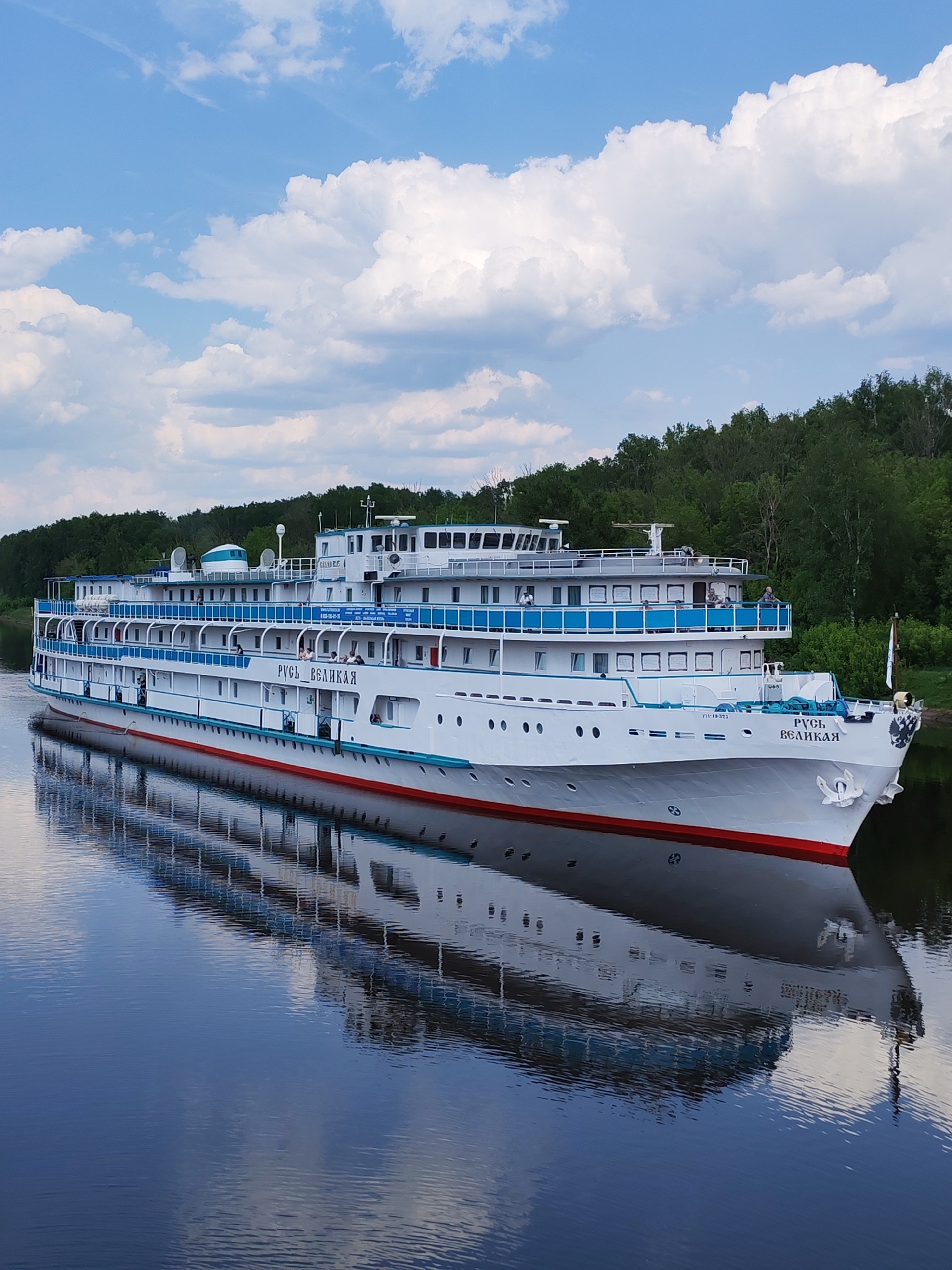 A large white river cruise ship, the "Rus Velikaya," is docked on a calm body of water, likely the Moscow Canal near Yakhroma, Russia, indicated by the surrounding green trees and the vessel's position on what appears to be a canal or wide river. The ship has multiple decks with blue railings and a red stripe along the waterline. The name of the ship is written in Cyrillic on the side. The sky above is partly cloudy with a mix of blue sky and white clouds. The water surface reflects the ship and the sky, creating a symmetrical effect. There are a few people visible on the decks of the ship.