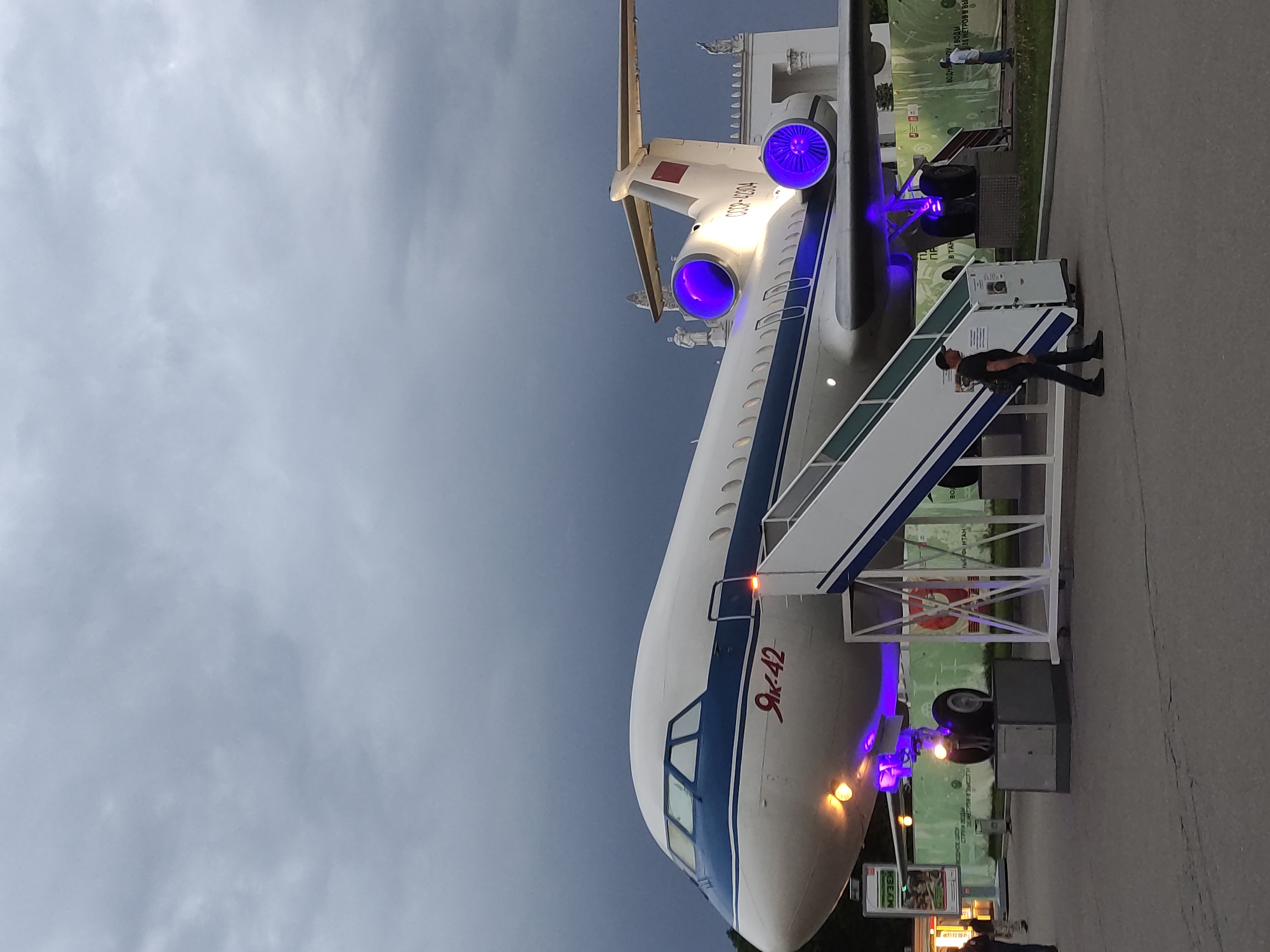 The image shows a Yak-42 passenger jet, with the text "Як-42" written on its side, and "CCCP-42304" on its tail. It's parked on a paved surface with a set of stairs attached to the entrance. A man is walking up the stairs. The plane is illuminated with blue lights near the engines and undercarriage. The sky is overcast. A sign with the text "Музей" is partially visible in the foreground.
Based on the architectural style visible in the background and the Cyrillic text on the plane, it is plausible this is located at the VDNKh (Exhibition of Achievements of National Economy) in Moscow, Russia, where numerous aircraft and other machinery from the Soviet era are on display.