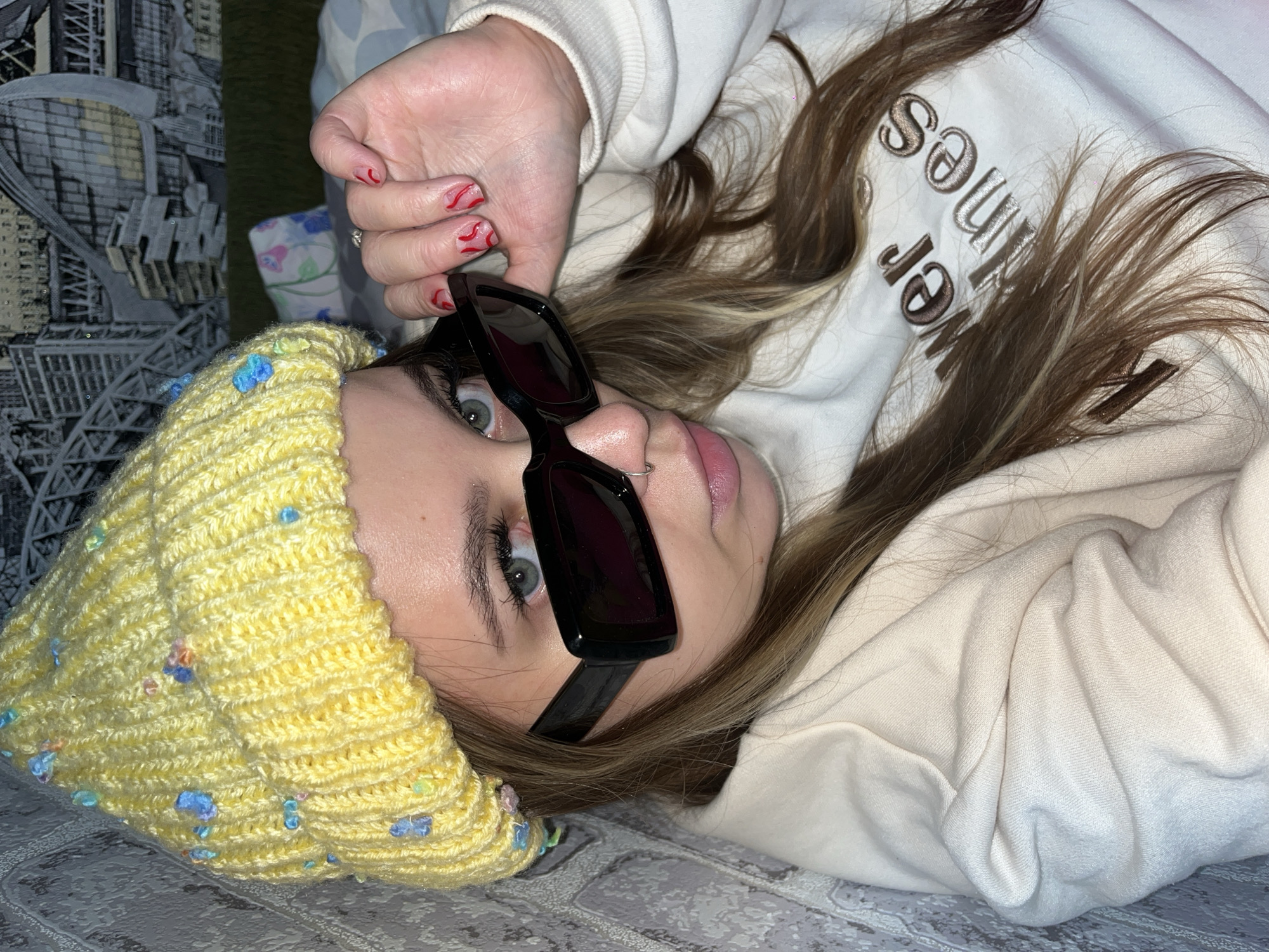 A young woman lies on her back, wearing a yellow knit hat with small blue and green decorative accents and black sunglasses. Her hair is light brown with blonde highlights, and she has a nose ring. She wears a white hooded sweatshirt, the hood up with "Sunflower" embroidered on the front. Her nails are painted with a red design. The background includes a textured gray surface and a wall or object with a black and white pattern. The city of Novopokrovskaya, Russia is not discernible in the scene.