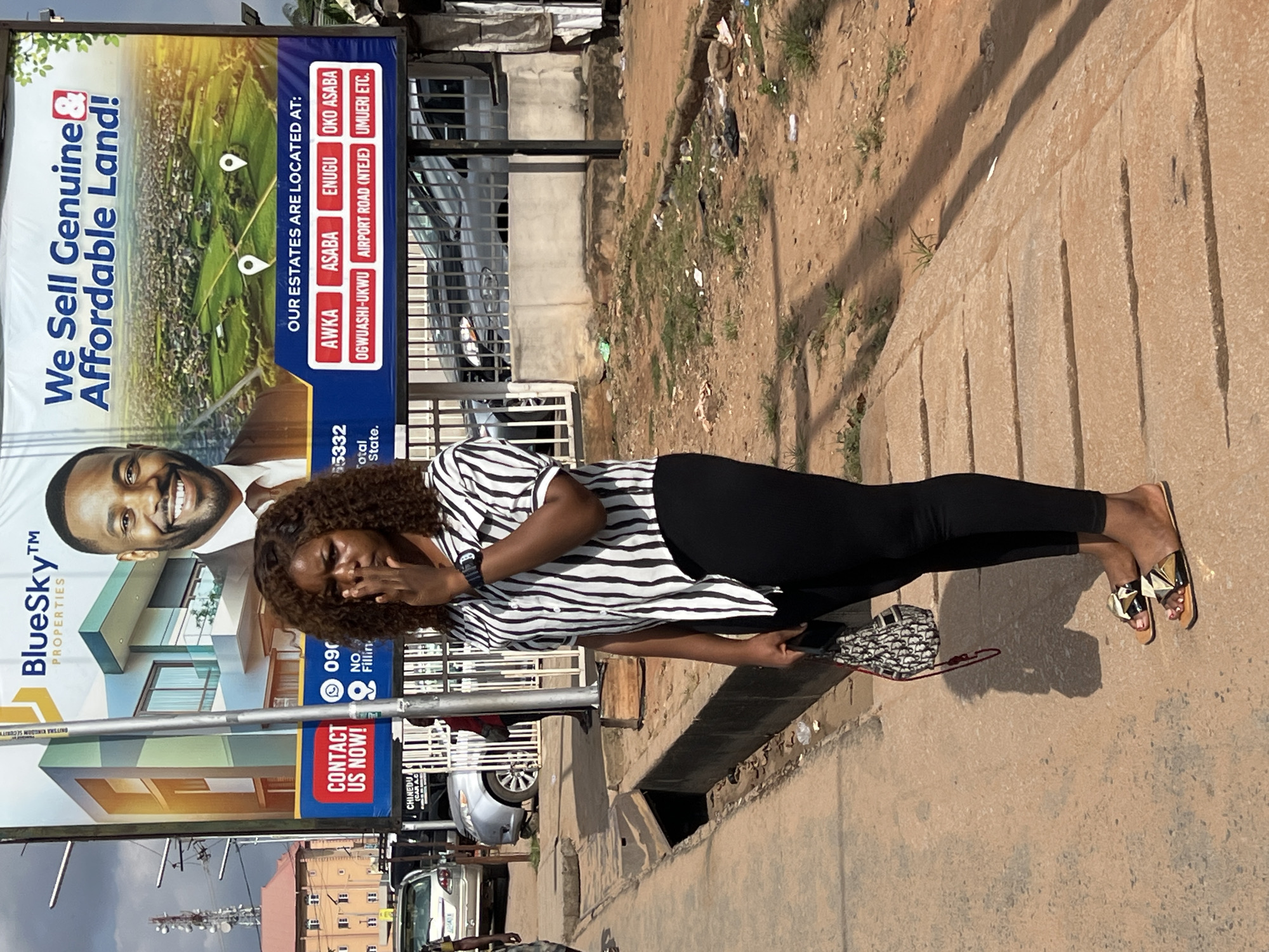 The image captures a bright outdoor scene in Onitsha, Nigeria, featuring a woman standing next to a large billboard on a sunny street. The woman, positioned on the right, has curly brown hair and is wearing a black and white striped short-sleeved top, black pants, and patterned flat sandals. She holds a smartphone in her left hand, and her right hand is partially covering her mouth, suggesting a smile or laugh as she looks towards the viewer. A small patterned handbag rests on the ground beside her feet.
A prominent billboard dominates the left side of the frame, advertising "BlueSky™ PROPERTIES" with the slogan "We Sell Genuine & Affordable Land!". The billboard features a smiling man and images of modern houses in a green landscape. It lists various estate locations, including "AWKA, ASABA, ENUGU, OKO ASABA, OGWUASHI-UKWU, AIRPORT ROAD (NTEJE), UMUERI ETC." Contact details like "CONTACT US NOW!" and partial phone numbers (e.g., "090 55332") are also visible, along with "ONITSIRA KINGDOM SECURITY" and "CHINEDU (CAR AC)".
The scene depicts an urban environment under clear skies, with strong sunlight casting distinct shadows. The ground consists of a concrete sidewalk next to a dusty area with sparse grass, and an open drainage ditch runs along the street. In the background, behind the billboard, other buildings, parked cars, and a utility pole with various antennas are visible, contributing to the impression of a bustling city street.