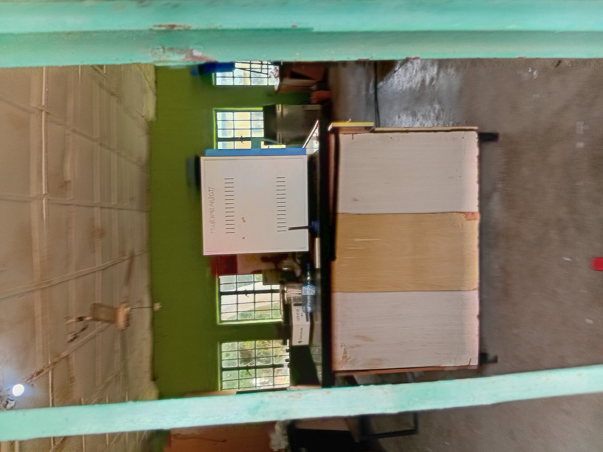 The image depicts an indoor scene, likely a laboratory or classroom, with a green painted wall. In the foreground, a wooden counter with three panels of varying shades of beige and light brown is visible. On the counter, a white apparatus with vertical lines is positioned, and next to it, a glass beaker and a box are present. Behind the counter, two windows with a grid-like pattern are seen, with trees and foliage visible through them, suggesting an outdoor environment beyond. A ceiling fan is mounted on the tiled ceiling, and a light fixture is also present. The floor appears to be concrete. Two thick, turquoise-colored vertical posts frame the scene on the left and right, obscuring parts of the room. The overall impression is that of a functional but perhaps somewhat dated or poorly maintained indoor space. Without additional context, it is impossible to pinpoint the exact location in Jalingo, Nigeria.
