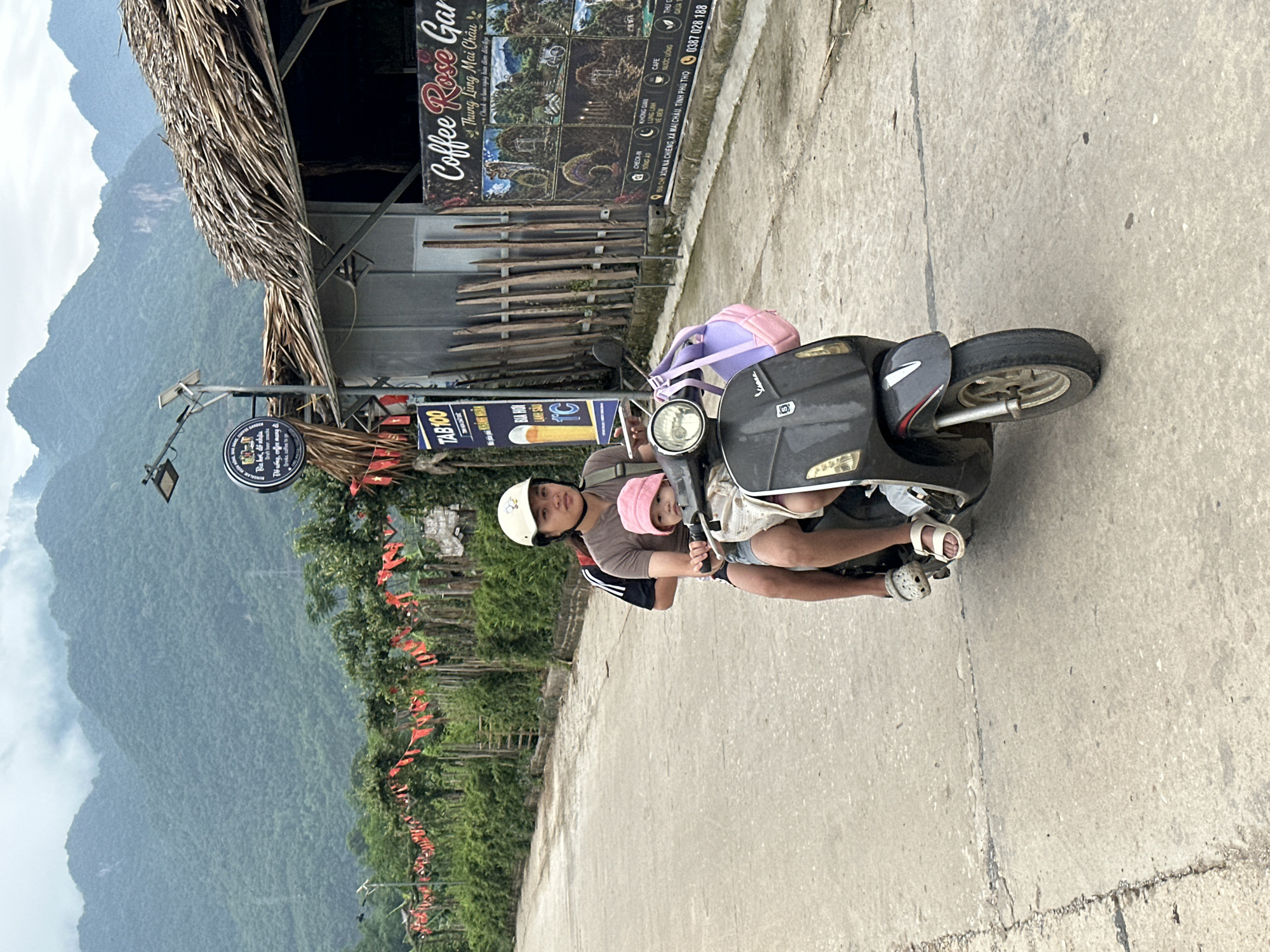 In Mai Chau, Vietnam, two individuals are riding a scooter on a paved road. The person in front, wearing a white helmet and a brown shirt, is operating the scooter. Behind them, a child wearing a pink hat is seated, also wearing a light-colored top. Both are looking forward. The scooter is dark gray with a white front fender and a silver trim. A purple and pink bag is attached to the back of the scooter. In the background, lush green mountains rise under a cloudy sky. A building with a thatched roof is visible to the right, adorned with red flags and signage, including one that reads "Coffee Rose Garden." The overall scene depicts a common mode of transportation in a rural Vietnamese setting.