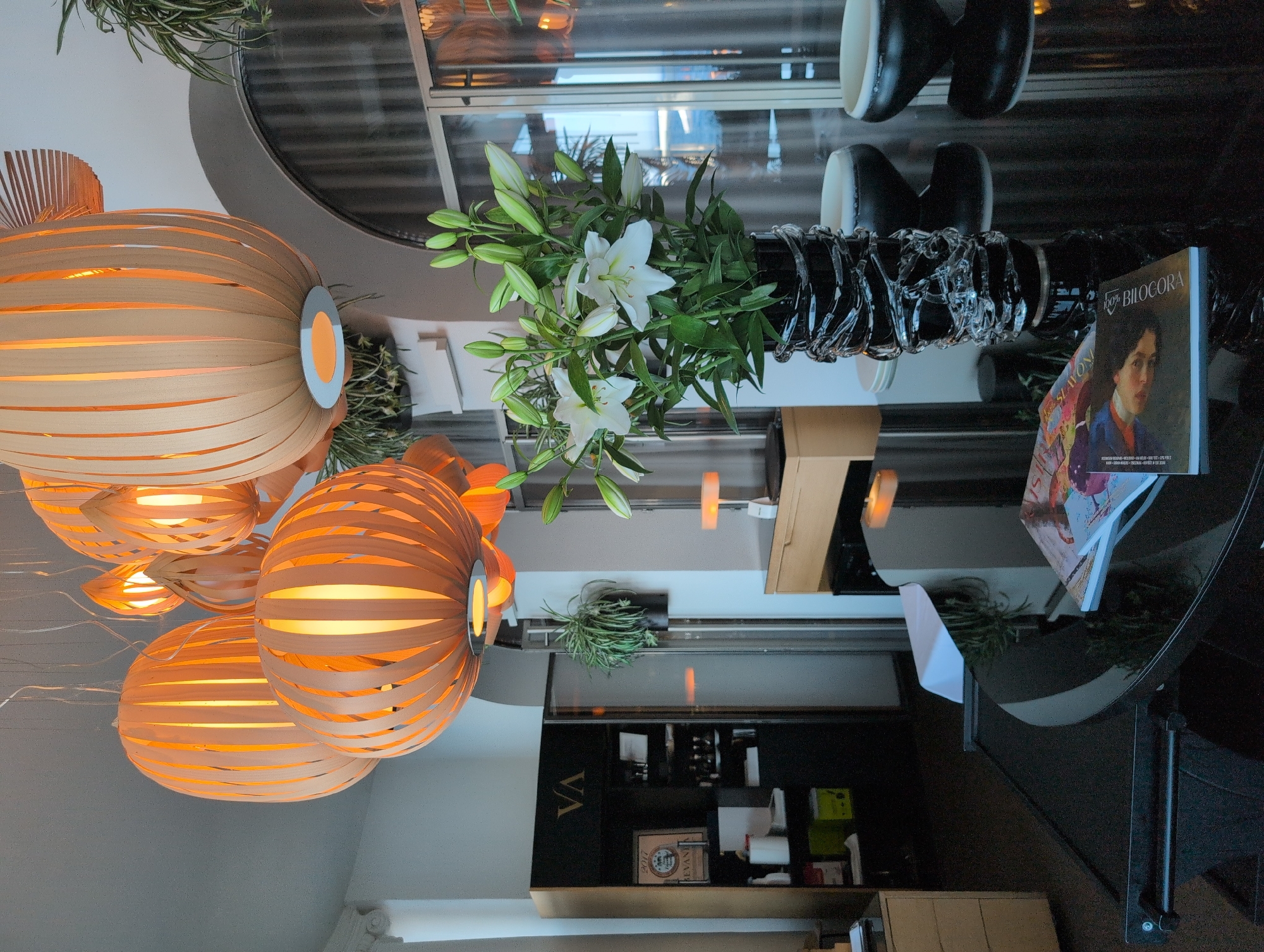 The image depicts an interior scene, possibly a restaurant or hotel lobby, with a focus on decorative elements and furniture. In the foreground, a glossy black table reflects the overhead lighting and displays two magazines. To the right, two dark, modern-style bowls or vases are visible. Dominating the central part of the image is a large black vase filled with a bouquet of white lilies and their buds.
The background features several large, pendant lamps made of light-colored wood strips, emitting a warm, orange glow. To the left, a black cabinet with glass doors, labeled with "VA", contains shelves stocked with various items, possibly beverages or decor. A potted plant hangs near the cabinet. Further back, a desk with a light wood finish is partially visible. The overall environment suggests a sophisticated and contemporary setting. There are no people visible in the image. The scene is well-lit, with the warm glow of the lamps creating an inviting atmosphere.
