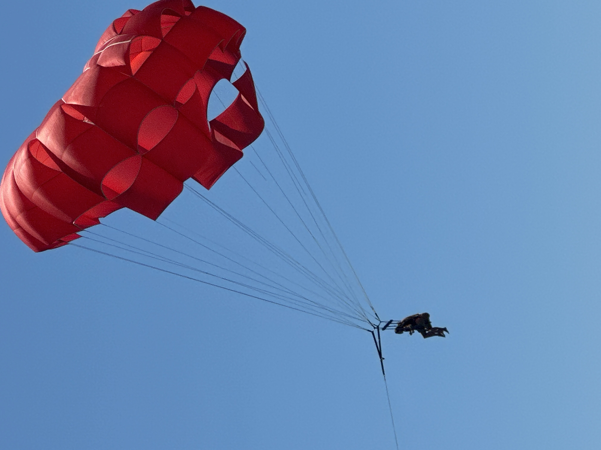 A person is parasailing against a clear blue sky. The parasail is bright red and the person is suspended beneath it in a harness. The scene is shot from a low angle, looking up at the parasail. The image does not provide any specific details about the location in Boreti, Montenegro.