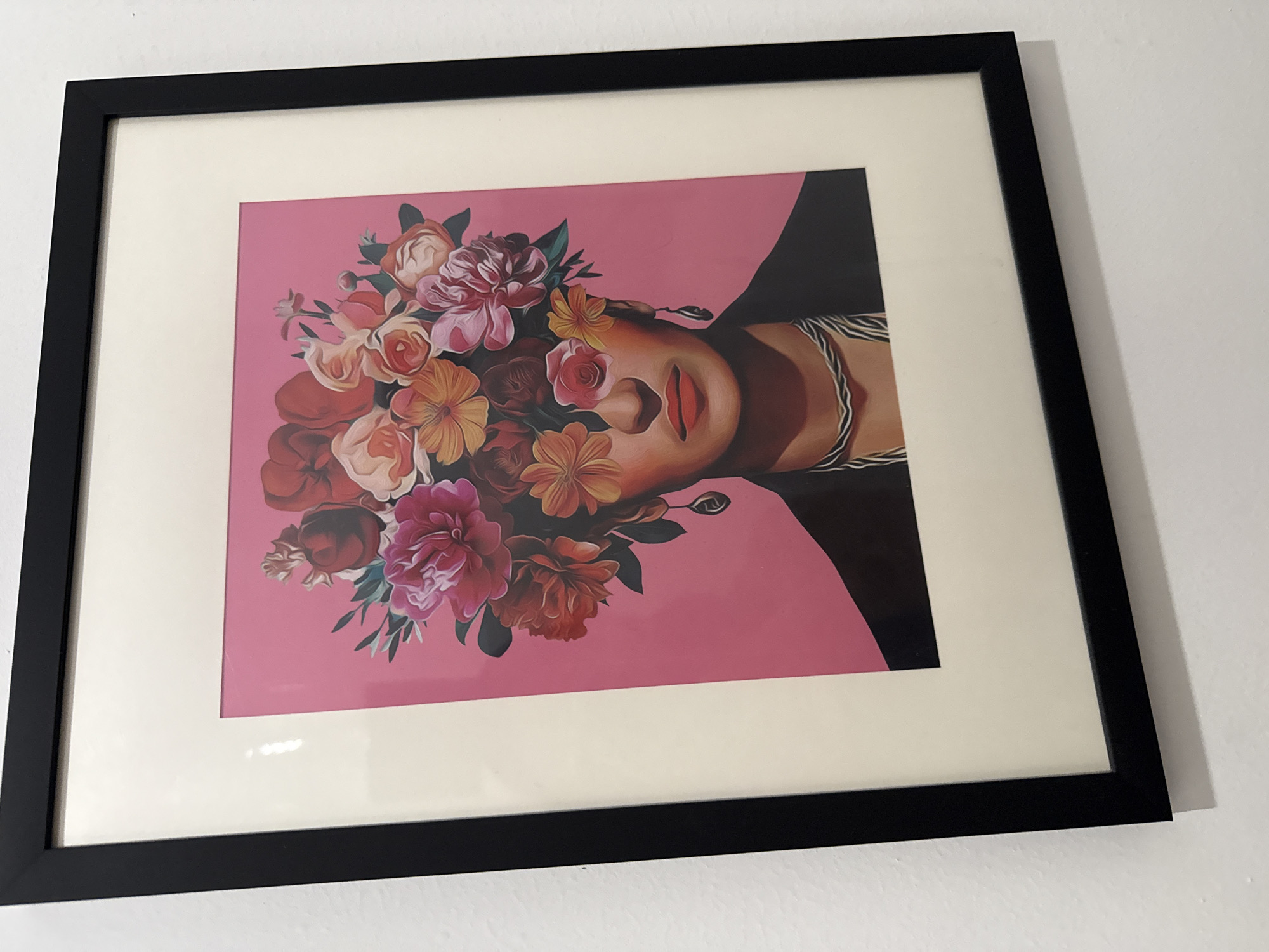 The image shows a framed print of a woman's face adorned with flowers, reminiscent of Frida Kahlo, against a pink background. The print is framed with a black frame and a wide, off-white mat. The frame is hanging on a white wall. The location is impossible to determine from the image.