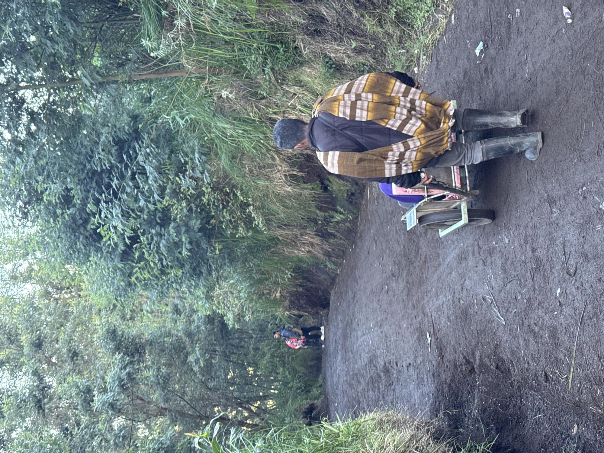 The image shows a man in black boots, grey pants, a black jacket, and a tan and white patterned shawl, sitting on a wheeled cart on a muddy path. The path is lined with dense green vegetation. Further down the path, two figures can be seen walking. The location is in an area with dense vegetation in Unknown, Indonesia.