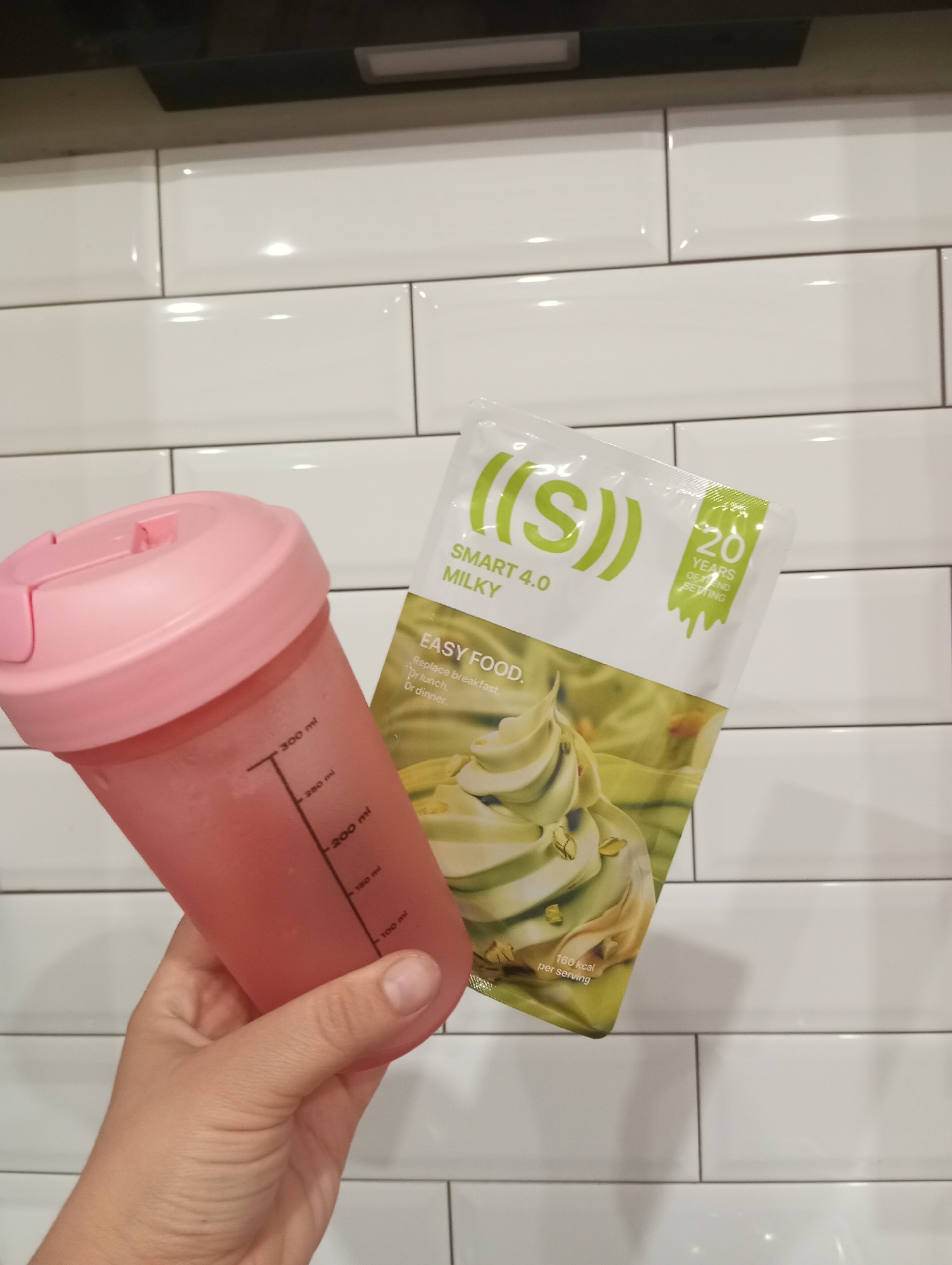 A hand holds a pink shaker bottle filled with a pink liquid and a package of "SMART 4.0 MILKY" easy food. The background is a tiled white wall. The bottle has measurement markings up to 300 ml. The package features a picture of green ice cream and text that reads "20 YEARS OF TREND SETTING" and "160 kcal per serving".