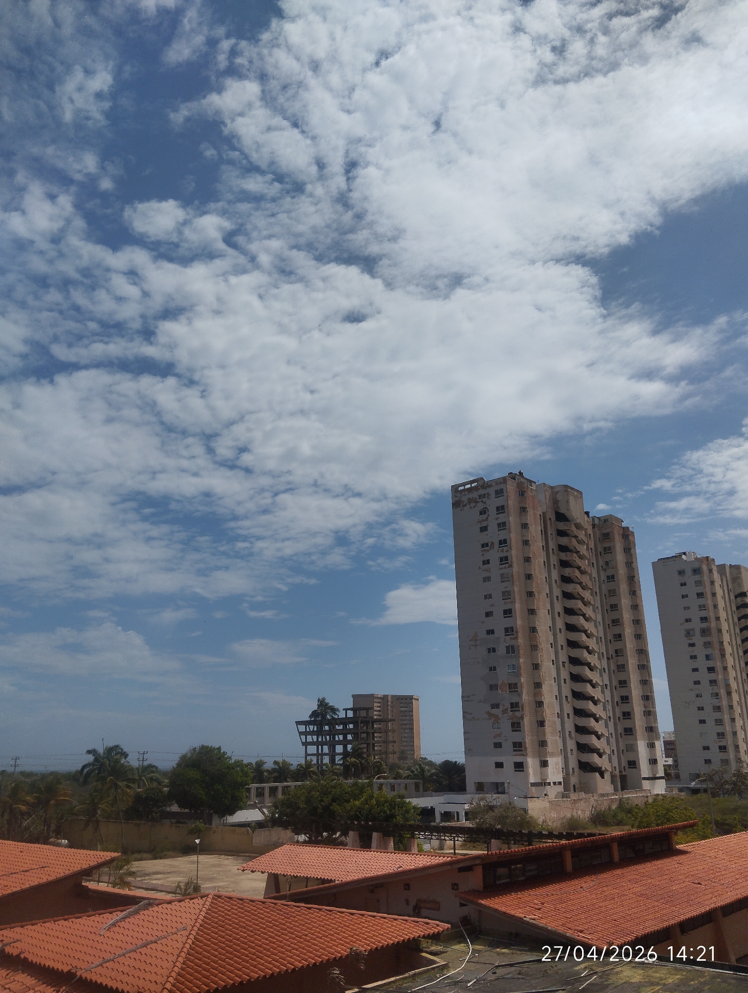 The image captures an outdoor scene in Porlamar, Venezuela, on a bright, partly cloudy day. The sky, filling the upper half of the frame, is a clear blue adorned with numerous scattered white cumulus clouds.
In the foreground, prominent terracotta-tiled roofs of several low-rise buildings are visible, appearing neat and well-maintained. The midground features a band of lush green vegetation, including several tall palm trees and other foliage, bordering an empty, unpaved lot where a single lamppost stands. A low wall or fence runs across this section.
The background is dominated by several tall, multi-story buildings. On the right, two large, weathered high-rise structures stand, characterized by stained facades, visible signs of disrepair, and external staircases or balconies. These buildings appear aged and possibly abandoned or neglected. Further to the left in the background, a distinct skeletal structure, resembling the framework of a building either under construction or in a state of advanced decay, is visible, alongside another beige-colored high-rise.
No people or specific activities are visible within the frame, contributing to a quiet, possibly desolate atmosphere. The scene contrasts the well-kept foreground roofs with the worn and neglected appearance of the background high-rises. A digital timestamp "27/04/2026 14:21" is present in the bottom right corner of the image.