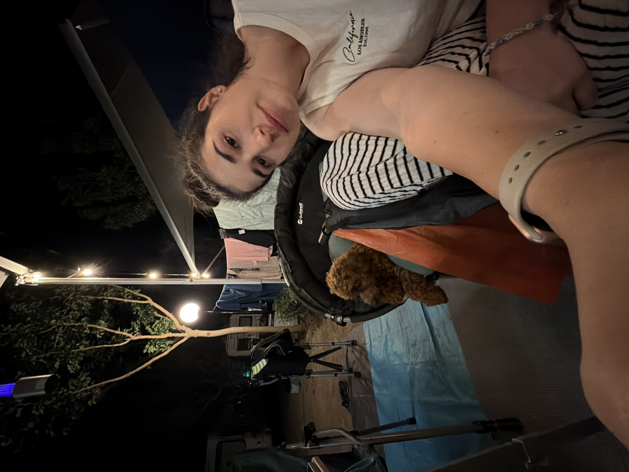 A low-angle, medium shot captures a woman and a small, brown poodle in an outdoor setting at night, likely a campground. The woman, in the foreground, is looking at the camera with her hair pulled up in a bun and is wearing a white t-shirt with the text "California Los Angeles Est. 1996". She is resting her arm on her lap, which is covered by black and white striped pants. A light-colored bracelet adorns her wrist. To her left, a poodle sits inside a dark-colored carrier. In the background, a tree with sparse leaves is illuminated by a white light at its base and a blue light fixture hanging from its branches. String lights are visible above, and laundry is hanging on a line behind the tree. Camping equipment, including chairs and what appears to be a camper van, are present. The ground is covered with a blue tarp and a grey mat. The overall scene suggests a relaxed evening at a campsite near Πόρτες, Greece.