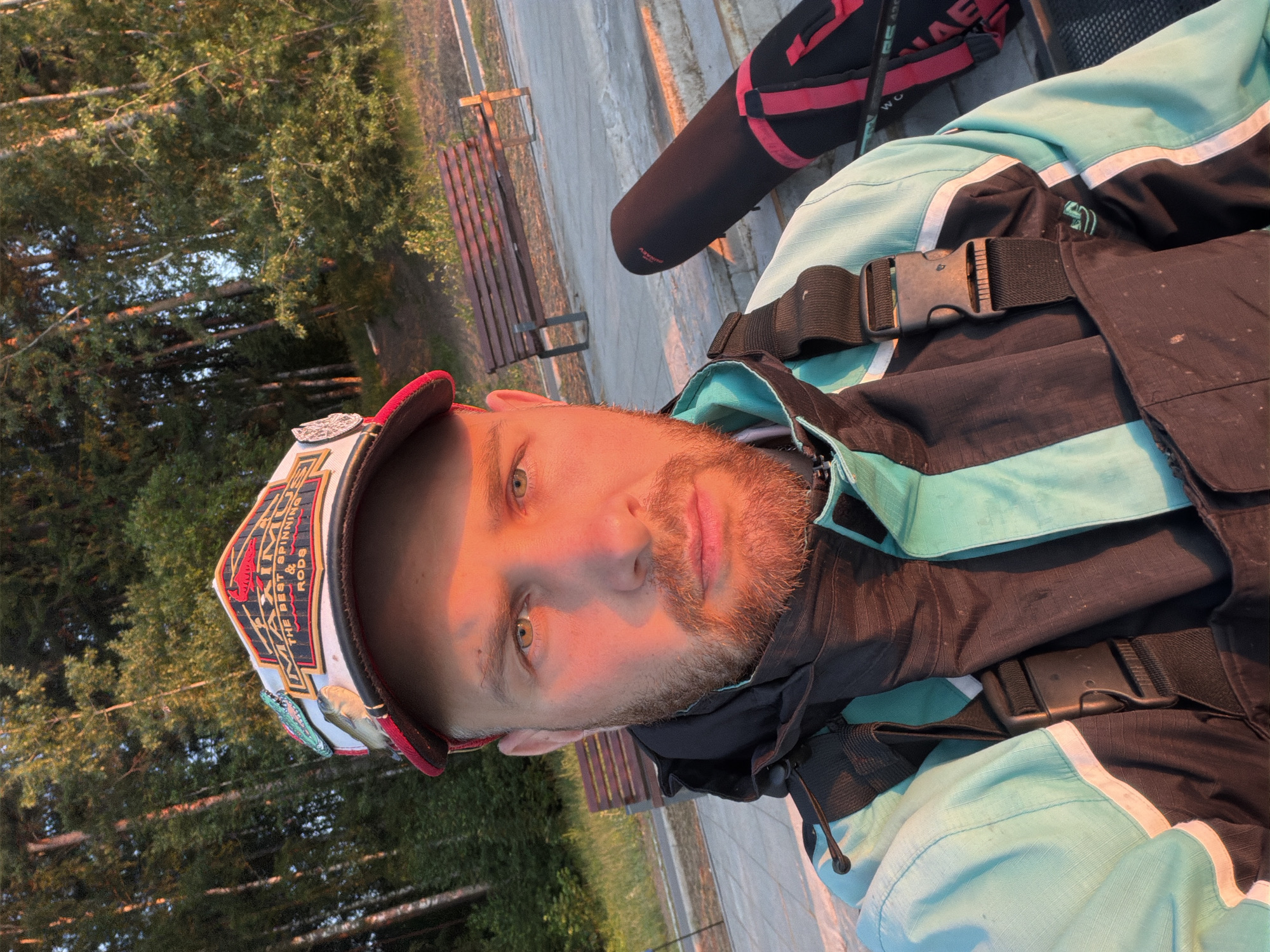 A man is taking a selfie outdoors. He is wearing a fishing vest and a cap with fishing-related decorations, including a small fish and an alligator. In the background, there are trees and a park bench. The lighting suggests it is either sunrise or sunset, casting a warm glow on the man's face. A fishing rod case is visible to the right of the man. The location appears to be a park or waterfront area in Chaikovsky, Russia, possibly near the Kama River given the foliage and open space.