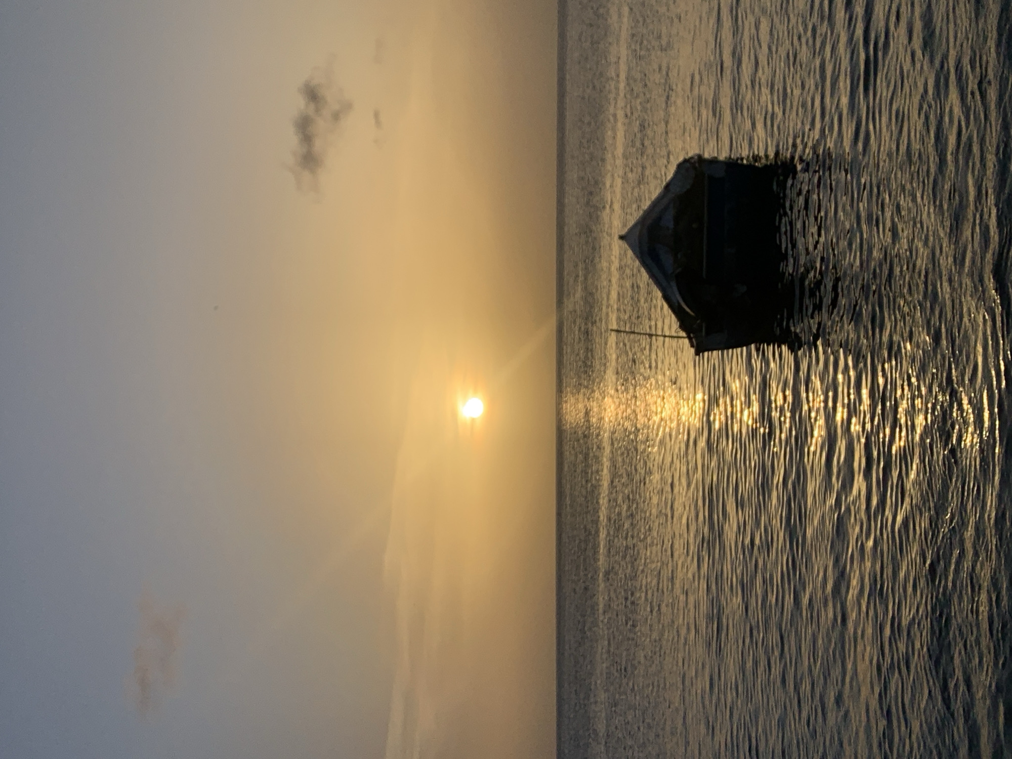 A lone, dark-hulled fishing boat drifts on calm, dark water under a pale orange sky, nearing sunset. The sun, a bright, luminous orb, is positioned slightly to the right of the frame's center, casting a shimmering golden path across the water's surface. Two small, wispy clouds are visible in the sky, one in the upper left quadrant and another in the upper right. The boat, silhouetted against the water and sky, appears to be empty and at rest. A single, thin pole extends upward from within the boat, possibly a mast or oar. The water is gently rippled, reflecting the warm hues of the sky. This scene captures the tranquil beauty of a sunset over the ocean, likely near the coast of Juan Griego, Venezuela, a popular location for fishing and enjoying coastal views.