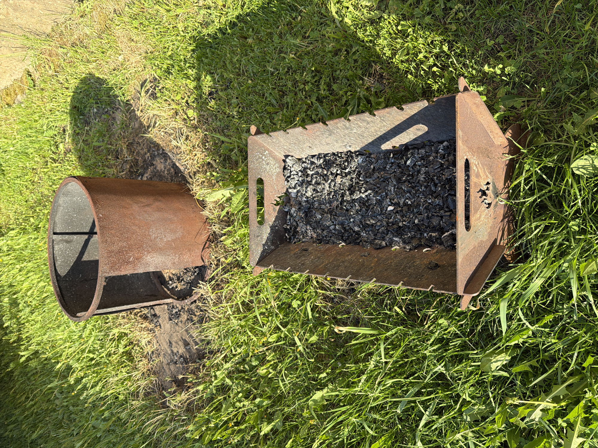 The image depicts a rusted, two-part metal charcoal grill on a patch of green grass. The first part is a cylindrical container with a stand; the other is a rectangular, cage-like structure filled with used charcoal or ash. The location is Bolsheustyikinskoye, Russia. No people or actions are visible. The environment is grassy and outdoors, with a small concrete edge visible in the top left corner.