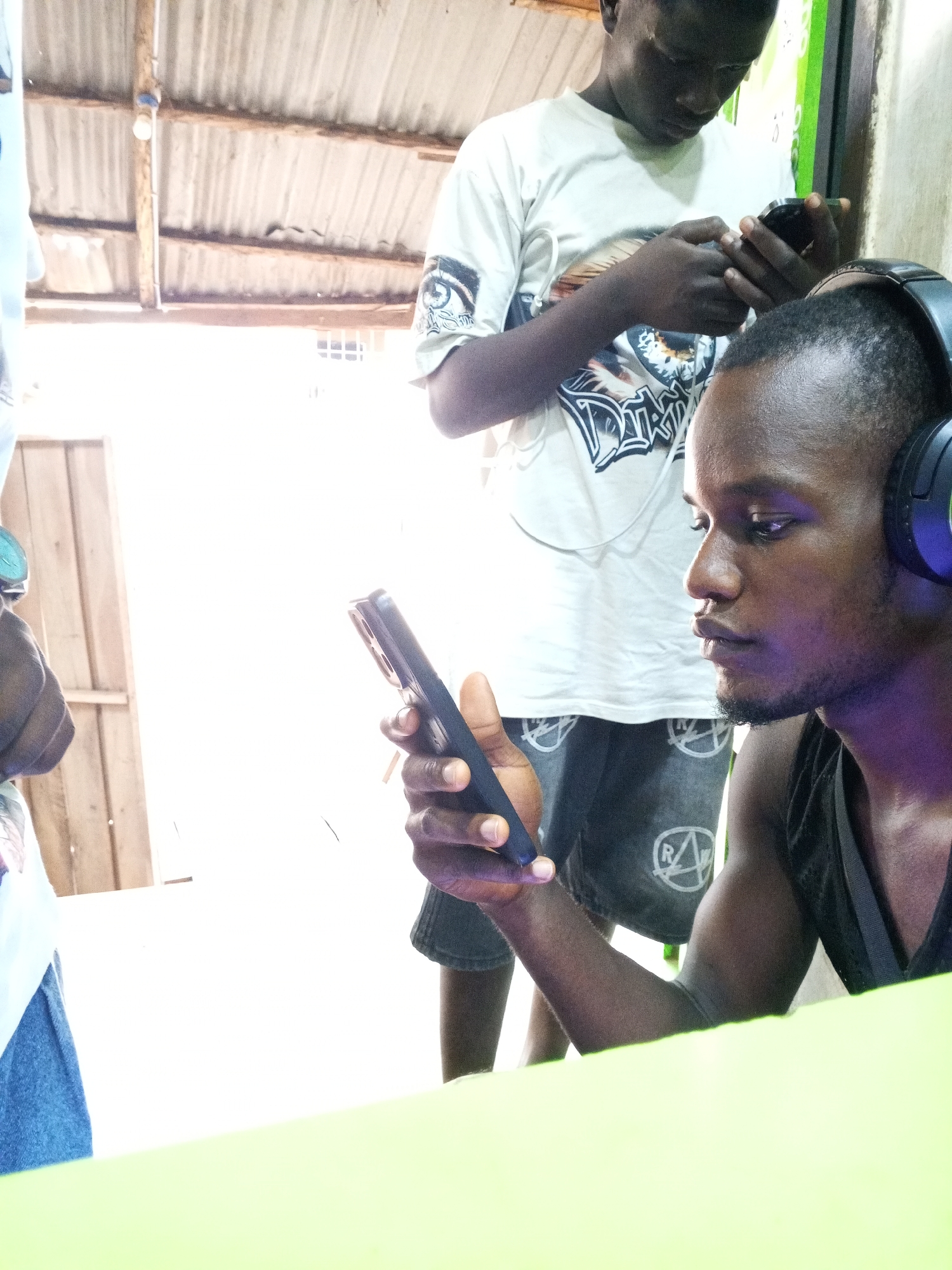 Two men are in an interior space. One man is in the foreground and wears headphones. He looks at a mobile phone in his hand. The second man stands behind him and is also looking at a mobile phone. The second man is wearing a t-shirt with a graphic print. Overhead is a corrugated metal roof structure. The foreground contains a lime green surface. The location is assumed to be Lokoja, Nigeria.