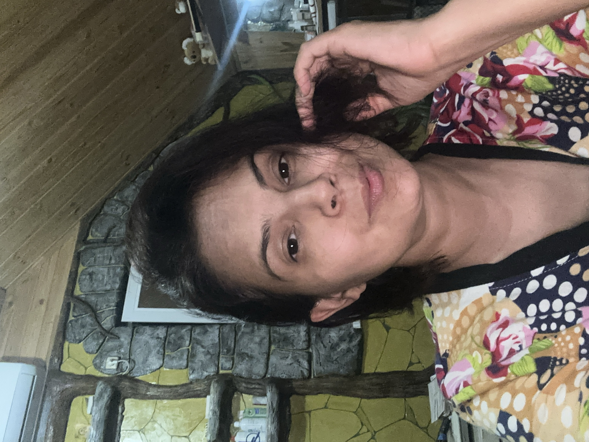 A woman is captured in a selfie, with her face filling most of the frame. She has dark hair, styled to fall around her face, and appears to be touching her hair with her right hand. Her eyes are open, and she has a slight smile on her lips. She is wearing a colorful top with a floral and polka dot pattern.
The background suggests an indoor setting with a rustic or unconventional design. To the left of the woman, a wall features a textured design resembling stone and wood, with shelves holding several bottles and containers. Above this, a white air conditioning unit is visible. The ceiling is made of wooden planks. To the right, there is a shelf with small decorative figures, possibly teddy bears, and other indistinct objects. The lighting suggests it is nighttime or the room is dimly lit, with a visible light source to the right creating a glare.
The provided information states the image's location is Kalininskaya, Russia. This detail is external to the visual content of the image itself and cannot be confirmed from the image alone.