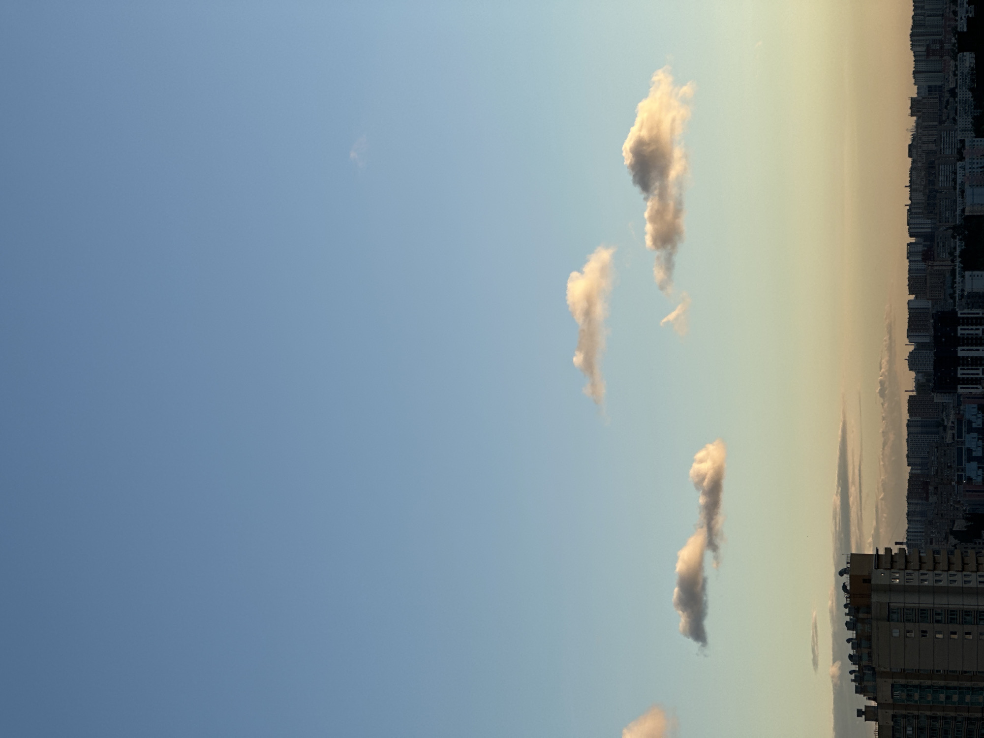 The image depicts a cityscape of Novosergievka, Russia, under a clear sky with scattered clouds. The lower half of the image shows a dense urban landscape with numerous multi-story buildings. A prominent, tall, light-colored building with visible balconies and a distinctive roofline is located on the left side of the frame. Behind it, a sprawling panorama of apartment blocks and other structures extends across the horizon.
The upper half of the image is dominated by the sky, which transitions from a light blue near the horizon to a deeper blue at the top. Several wispy, light-colored clouds are dispersed across the sky, with some catching the warm light of either sunrise or sunset. The lighting suggests that the photograph was taken during the golden hour. There are no visible people or vehicles in the image, focusing entirely on the architecture and the sky. The environment appears to be an urban setting, likely a residential area, with a clear view of the sky above.
