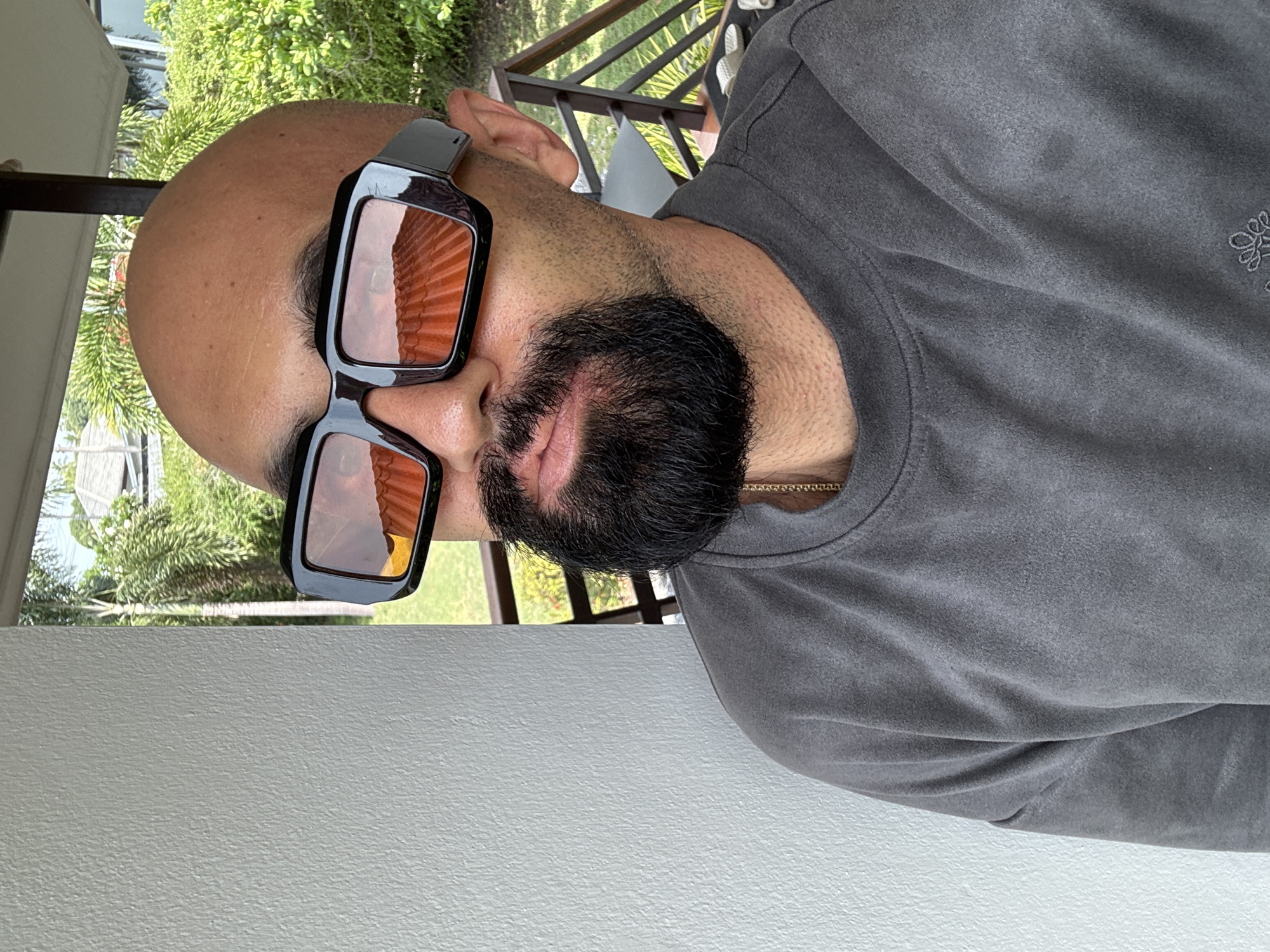The image shows a close-up, head-and-shoulders shot of a bald man with a beard, wearing large, dark-framed sunglasses with orange lenses. He is wearing a dark gray t-shirt with a visible gold chain around his neck. The background suggests an outdoor tropical setting, with lush green foliage, palm trees, and a glimpse of buildings with red tile roofs, indicative of a resort or residential area in Ko Samui District, Thailand. The man is positioned in front of a textured white wall on the left, and the right side of the image shows a wooden railing and more greenery, suggesting a balcony or patio. The lighting is bright, suggesting daytime.