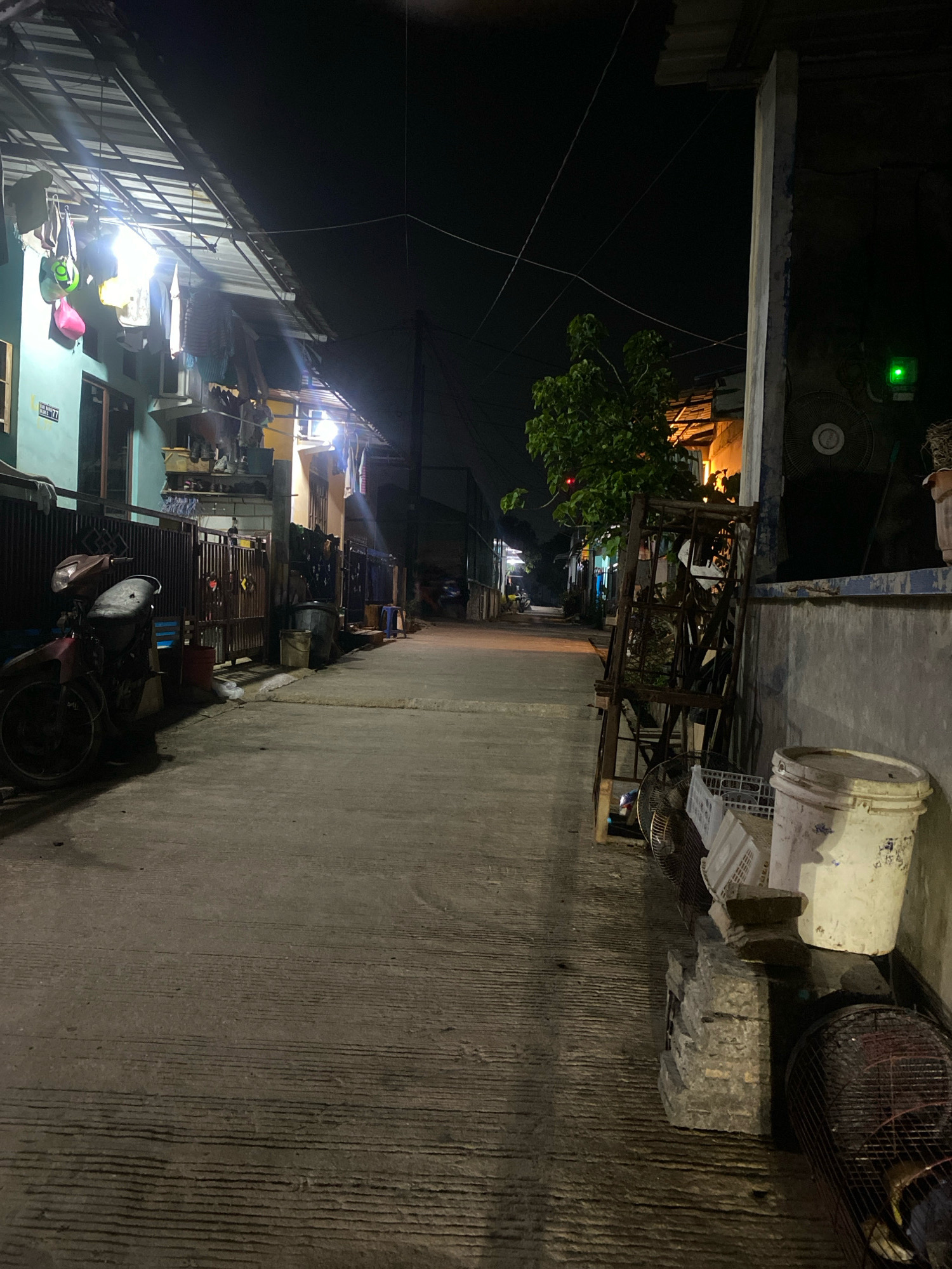 This is a low-light, eye-level, full shot of a narrow, paved alleyway at night. The alley is lined with residential buildings and fences. On the left, a motorcycle is parked in front of a metal fence. Clothes are hanging from a line under a covered awning. Lights illuminate the area in front of the buildings, casting long shadows. On the right side of the alley, there are various objects stacked near a wall, including buckets, fans, and what appears to be a metal shelving unit. A large tree with green foliage is visible in the middle distance on the right. The alleyway extends into the distance, with more buildings and faint lights visible. The overall impression is of a modest, urban residential area at night.