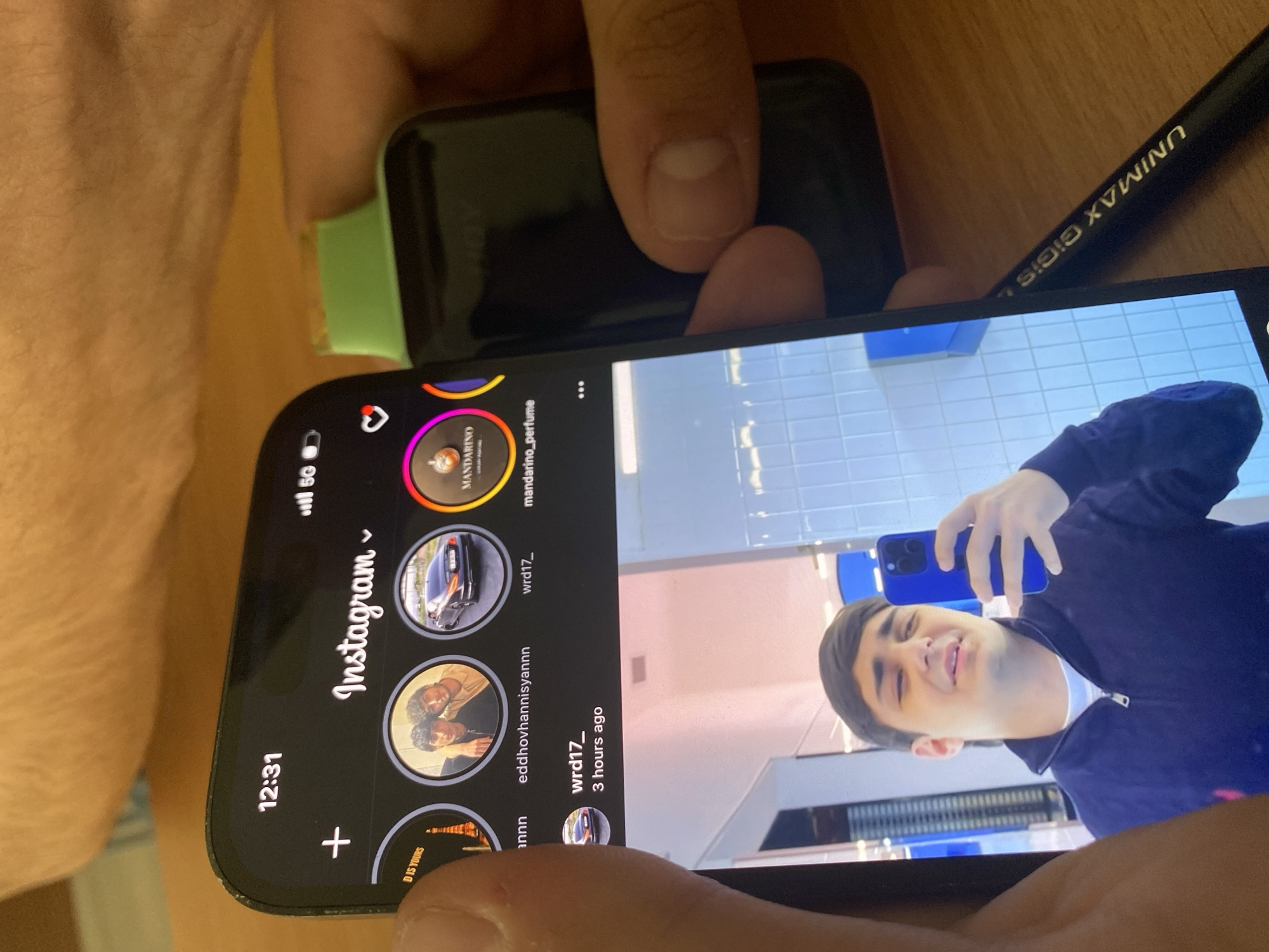 The image captures a close-up, rotated view of a person's hands holding a black smartphone, with a smartwatch featuring a light green band visible on their wrist. A black pen with "UNIMAX GIGIS" inscribed on it rests on a wooden surface in the background.
The smartphone screen displays the Instagram application. The time shown on the phone is "12:31", along with a "5G" network indicator. The Instagram interface shows several story circles from accounts such as "MANDARINO", "mandarino_perfume", "wrd17_", and "eddhovhannisyannn", with one story containing the text "D IS YOURS".
The central focus of the screen is an Instagram post or story from "wrd17_", uploaded "3 hours ago". This content features a young male taking a selfie or being photographed, holding a blue smartphone. He wears a dark blue zip-up top over a white t-shirt and displays a slight smile or smirk. The background of his image reveals a light blue tiled wall, suggesting an indoor setting with possibly reflective surfaces, such as a modern restroom or changing area. The bright lighting on the screen indicates it is daytime. The overall scene depicts casual engagement with social media in Yerevan, Armenia.