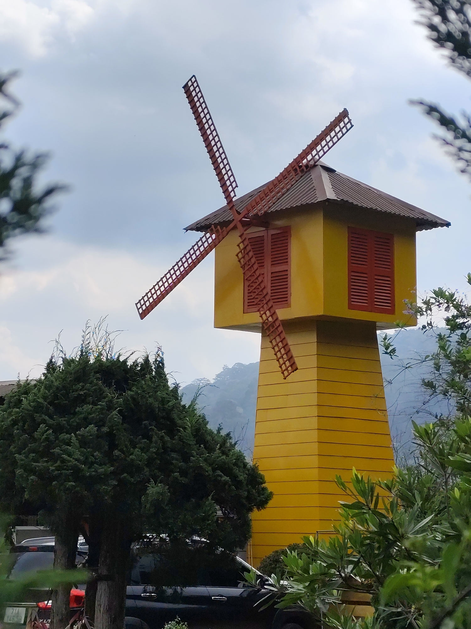 This outdoor daytime image captures a vibrant yellow decorative windmill structure, set against an overcast sky and hazy mountainous backdrop in Da Lat, Vietnam. The windmill features a tall, octagonal yellow base that tapers upwards, topped by a box-like yellow section with red louvered windows on its visible sides. A reddish-brown corrugated roof covers this upper section. Four prominent reddish-brown wooden blades, designed with a grid pattern, extend from the top, fixed in a stationary position.
The windmill is framed by lush green vegetation. A large, dense conifer-like tree with dark green foliage stands to the left of the windmill, partially obscuring the lower part of the structure. Other green bushes and trees are visible in the foreground and around the base, some appearing blurred, creating a sense of depth.
Behind the large tree and at the base of the windmill, parts of dark-colored vehicles are visible, suggesting a parking area or roadside location. A black car is partially seen, and on its license plate, the text "49A 146.9" is discernable. The sky is largely covered by white and light gray clouds, indicating cloudy weather and providing diffused light. No specific activity or people are depicted; the scene is static and serene, highlighting the whimsical architecture amidst the natural scenery.