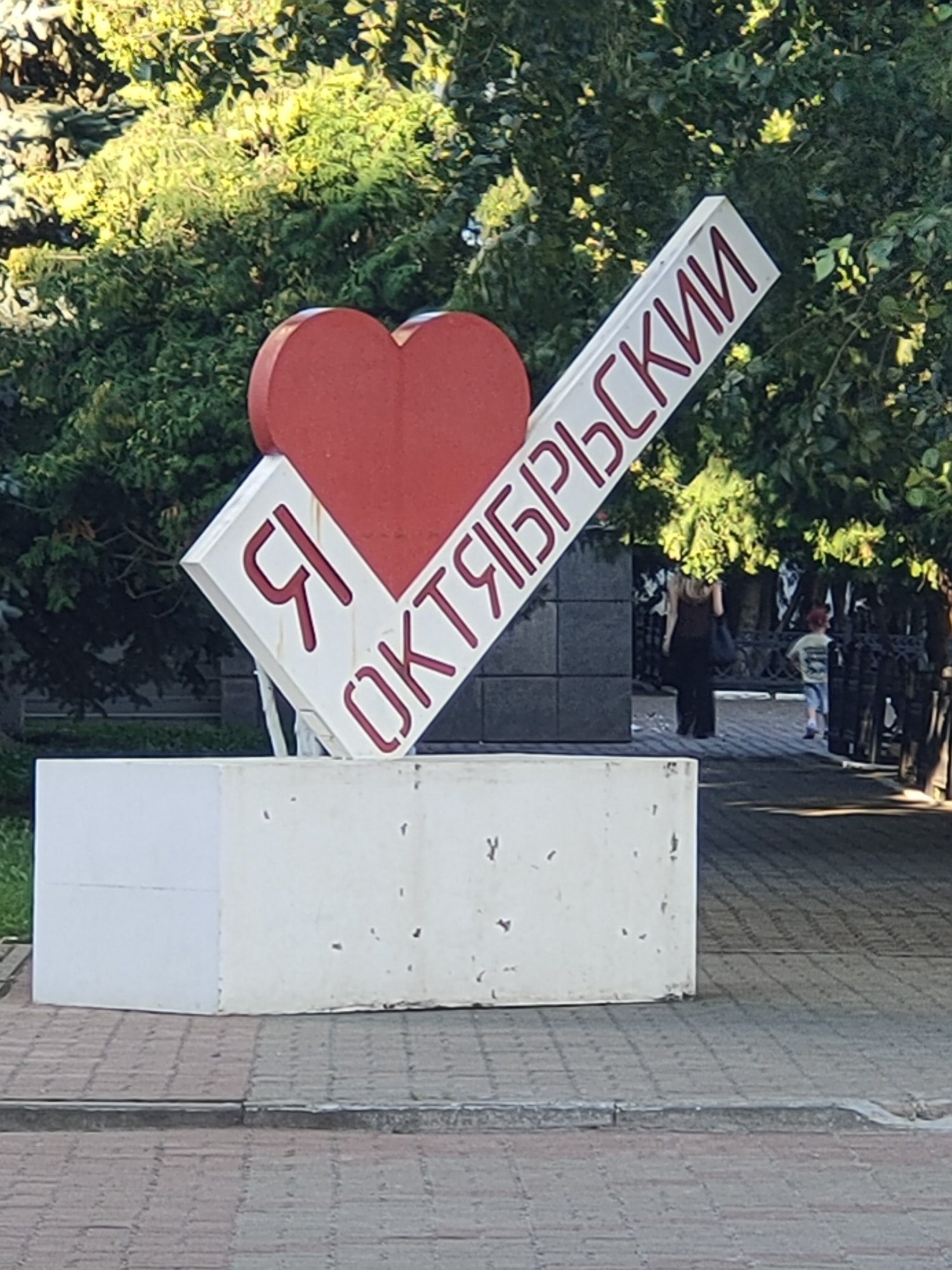 This is a full-shot, eye-level, outdoor photograph of a monument that reads "Я люблю Октябрьский" (I love Oktyabrsky) in the city of Oktyabrsky, Russia. The monument is a large, white, angular structure with a red heart shape on the left side. Red lettering spells out the phrase on the monument. Behind the monument, there are lush green trees and foliage. In the background, a woman with long blonde hair and a dark top and pants is walking away from the camera. A young child in a light-colored t-shirt and blue shorts is walking towards the camera on the right side of the image, behind a dark fence. The ground in front of the monument is paved with brickwork. The monument appears to be situated on a raised concrete platform.