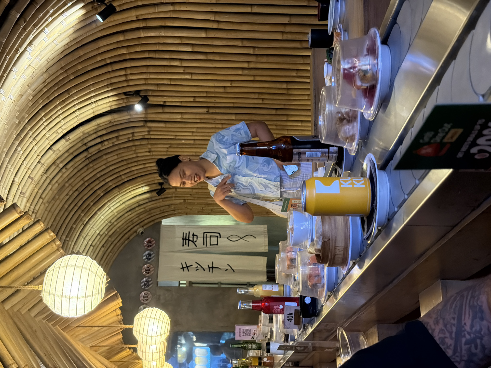 The image depicts the interior of a restaurant, likely a sushi bar, with a distinctive bamboo ceiling that curves overhead. The overall setting is dimly lit, with warm, ambient light emanating from paper lanterns hanging from the bamboo structure. The restaurant appears to be a conveyor belt sushi establishment, as evidenced by the stainless steel track with numerous small plates of food, each covered by a clear plastic dome, moving along it.
In the foreground, a person wearing a blue and white patterned shirt is partially visible, leaning towards the conveyor belt. They are smiling and pointing, suggesting engagement or interaction with the food or another person. A bottle of what appears to be beer is near them, along with a yellow can.
Further down the conveyor belt, various bottles of beverages are visible, including a red bottle with a label that reads "40K". In the background, a white banner with Japanese characters and a fish symbol hangs on a wall. The decor suggests a modern, possibly trendy Asian-inspired restaurant. There are some circular signs on the wall with numbers like "30K", "35K", "40K", and "50K", which could represent prices. The presence of multiple conveyor belts and the variety of food options imply a busy and dynamic dining environment. The overall atmosphere seems casual and inviting.