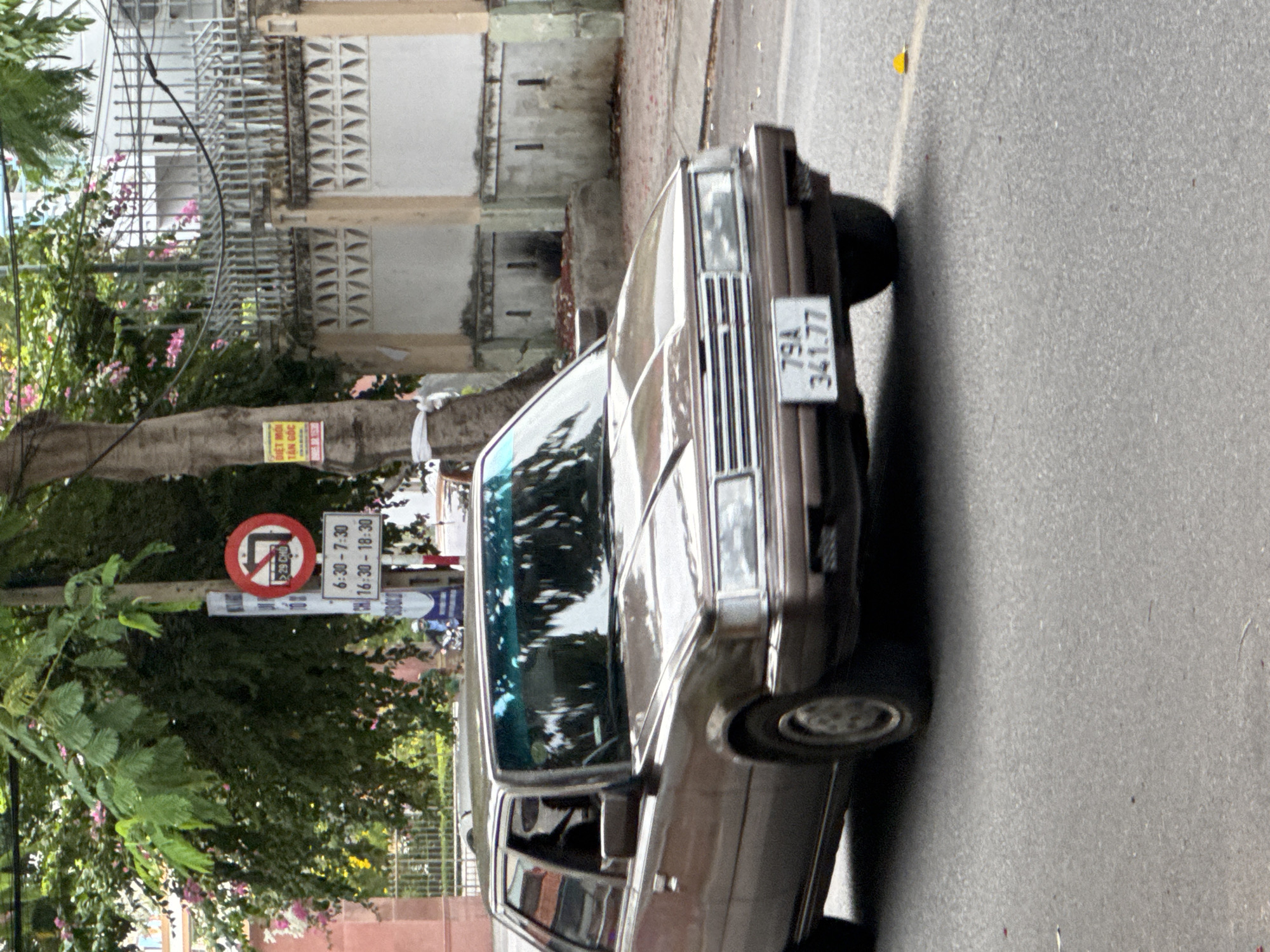 This is a medium shot, eye-level, outdoor, natural light photograph captured on a street in Nha Trang, Vietnam. The central subject is a brown, 1980s sedan, possibly a Toyota Camry or similar model, driving on asphalt. The car is seen from its front three-quarter angle, with the driver, a person with dark hair, visible in the driver's seat. The car's license plate reads "79A 341.77".
In the background, a tree with green foliage and pink flowers stands on the left. To the right of the tree, a traffic sign is mounted on a pole. This sign consists of a circular red prohibition symbol with a white arrow indicating a right turn into a T-junction, and below it, a white rectangular sign with black text. The text on the rectangular sign appears to be a time restriction, with "6:30 - 7:30" and "16:30 - 18:30". Adjacent to this sign, further to the right and partially obscured by the tree, is a yellow rectangular sign with black text.
Further into the background, a white metal fence runs horizontally across the image, separating the street from buildings. The buildings are light-colored with architectural details. A person on a motorcycle, wearing a blue shirt and a white helmet, is visible on the street behind the car, moving in the same direction. The overall environment suggests a typical urban street scene in Vietnam.