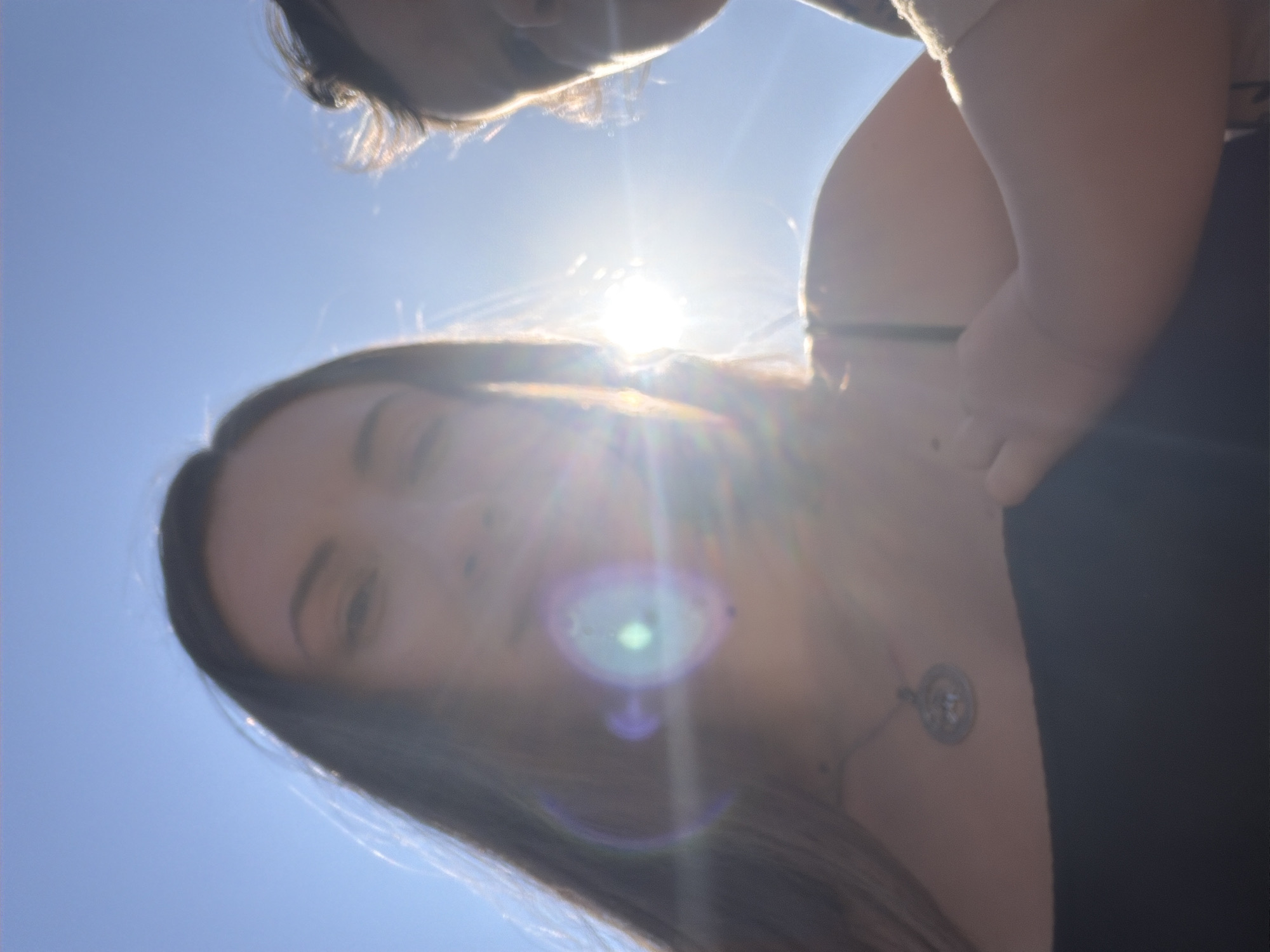 The image captures a close-up, low-angle selfie of two individuals against a bright blue sky, possibly in Dahab, Egypt, with the sun creating lens flare and obscuring details. A woman's face, in the foreground and slightly out of focus, is illuminated by the sun, with her dark hair framing her face. She wears a dark top and a pendant necklace. To her right, a child's face is partially visible, also backlit by the sun. The bright sunlight suggests an outdoor setting during the daytime.