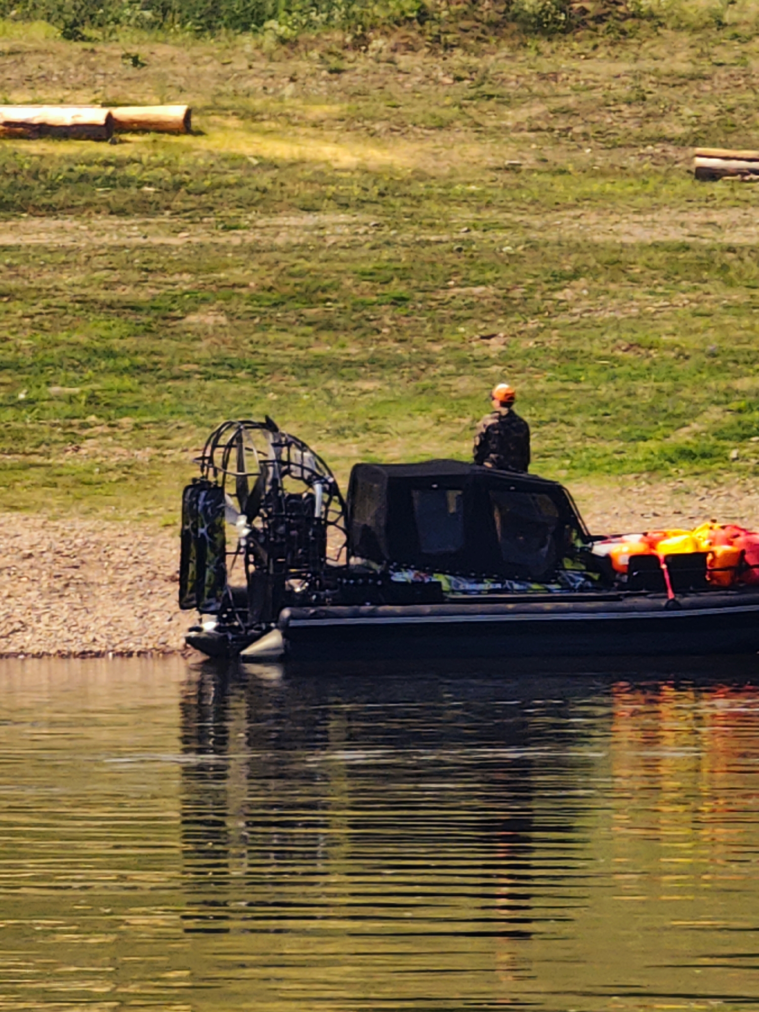 An airboat with a fan engine is docked on the shore of a body of water. A person wearing a camouflage jacket and an orange hat is standing on the back of the airboat. Several bright orange and yellow life jackets are stacked on the rear deck of the boat. The background consists of a grassy slope with some fallen logs. The water in the foreground is rippled, reflecting the boat and the surrounding environment. The image appears to be taken in Pogodaevo, Russia.