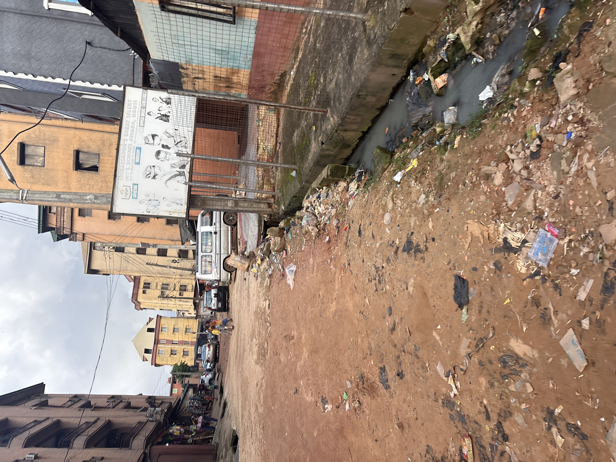 The image depicts a street scene in Onitsha, Nigeria, characterized by urban decay and a lack of sanitation. The foreground is dominated by a dirt road littered with various types of debris, including plastic bags, wrappers, and discarded paper. A murky, open drain runs along the right side of the road, also filled with trash and stagnant water.
In the mid-ground, several vehicles are present, including a white minivan and a silver sedan. A few individuals, seemingly children, are walking on the road. Buildings line both sides of the street, some appearing to be residential or commercial properties with multiple stories. One building features a large billboard advertising "Ambassador's Great Talent Int'l School Awada, Obosi," showcasing images of students in uniform.
The overall environment suggests an area experiencing significant neglect, with visible pollution and a general air of disrepair. The sky is overcast, contributing to a somewhat somber atmosphere. The buildings display a mix of architectural styles and conditions, with some showing signs of wear and tear. The street appears to be a public thoroughfare with moderate activity.