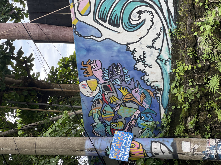A vibrant mural displays stylized sea creatures in a dynamic composition against a blue background with a white wave at the top. Various fish, including sharks and swordfish, are clustered together. The mural is affixed to a textured wall covered in foliage. In the foreground, a gray pole with a blue business card advertising "Vila Carita" is visible. Wires and tree branches with green leaves frame the image on the left.