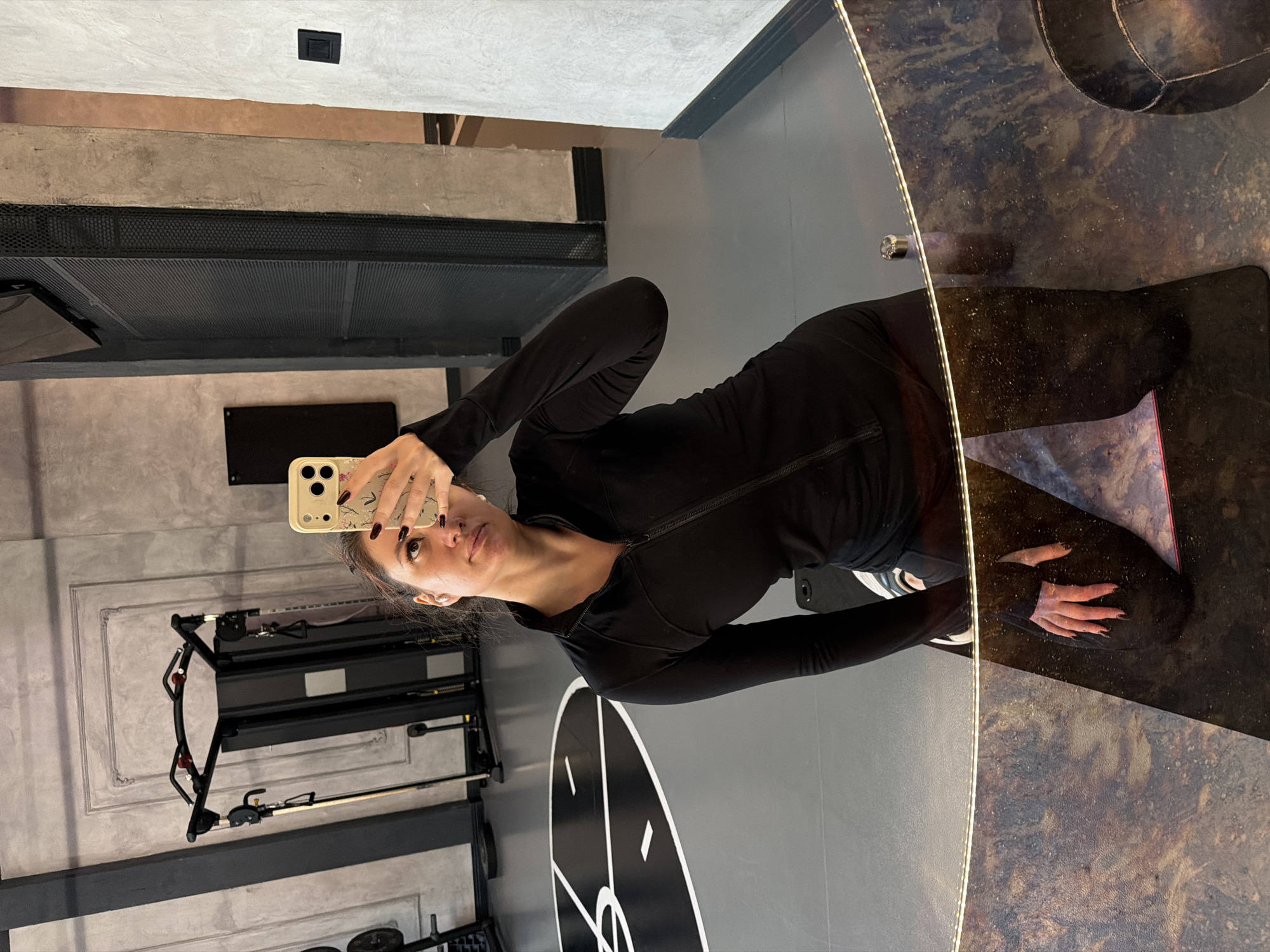 A woman is taking a mirror selfie in what appears to be a gym. She is wearing black athletic clothing. In the background, there is workout equipment, and the floor is gray with a white and black design. The gym's interior features gray walls and ceilings, with some sections showing exposed concrete. The mirror has an illuminated edge. It is not possible to determine the location in Belgrade, Serbia from the image.