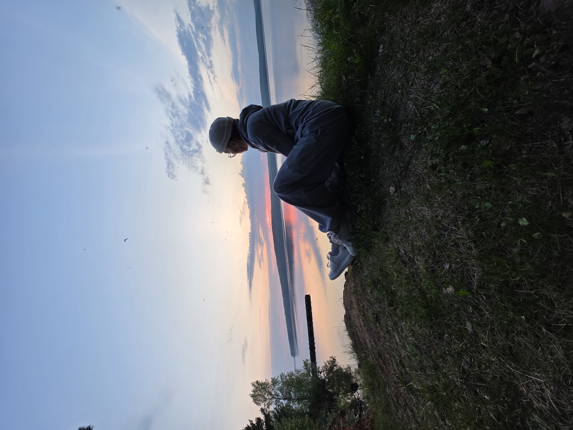 A person in a gray sweatsuit, wearing a beanie and glasses, crouches at the edge of a grassy embankment. The person is looking down, though the object of their attention is not visible. Behind them, the sun sets over a body of water, reflecting orange and pink hues. Trees and a dark structure are visible along the water's edge. The photograph appears to be taken in Chaikovsky, Russia, based on the general landscape and vegetation.