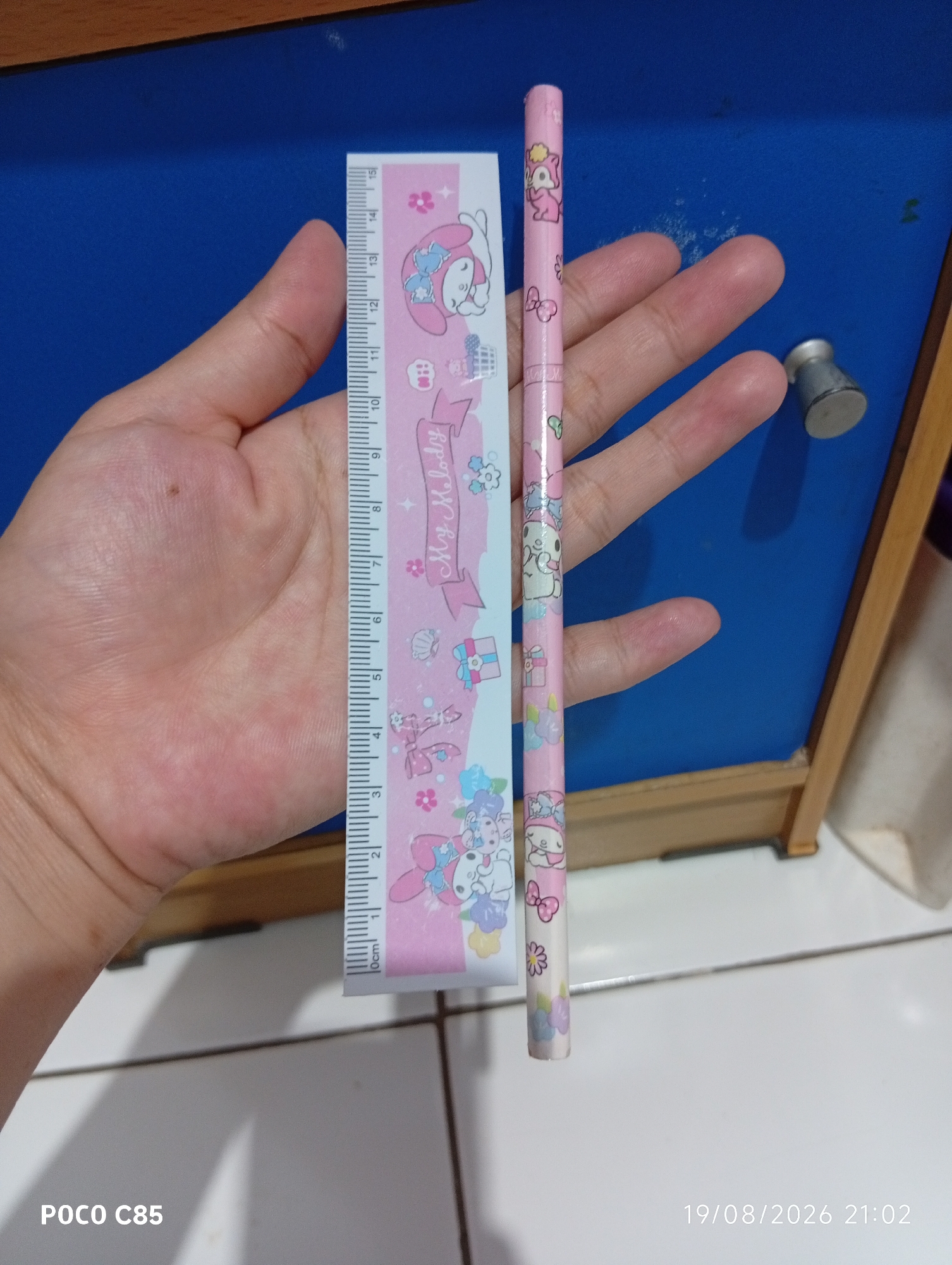 An adult's left hand holds a pink ruler with My Melody characters and a pink pencil with similar decorations. The ruler is marked in centimeters, starting from 0cm. The pencil is positioned parallel to the ruler. Both items are set against a blue surface with a wooden frame. A metallic cylindrical object is visible in the background, to the right of the hand. The foreground shows a white tiled floor. The timestamp "19/08/2026 21:02" and the text "POCO C85" are present in the lower portion of the image, indicating the date, time, and the device used for capture. The scene appears to be indoors, possibly in a room or on a desk, in South Tangerang, Indonesia.