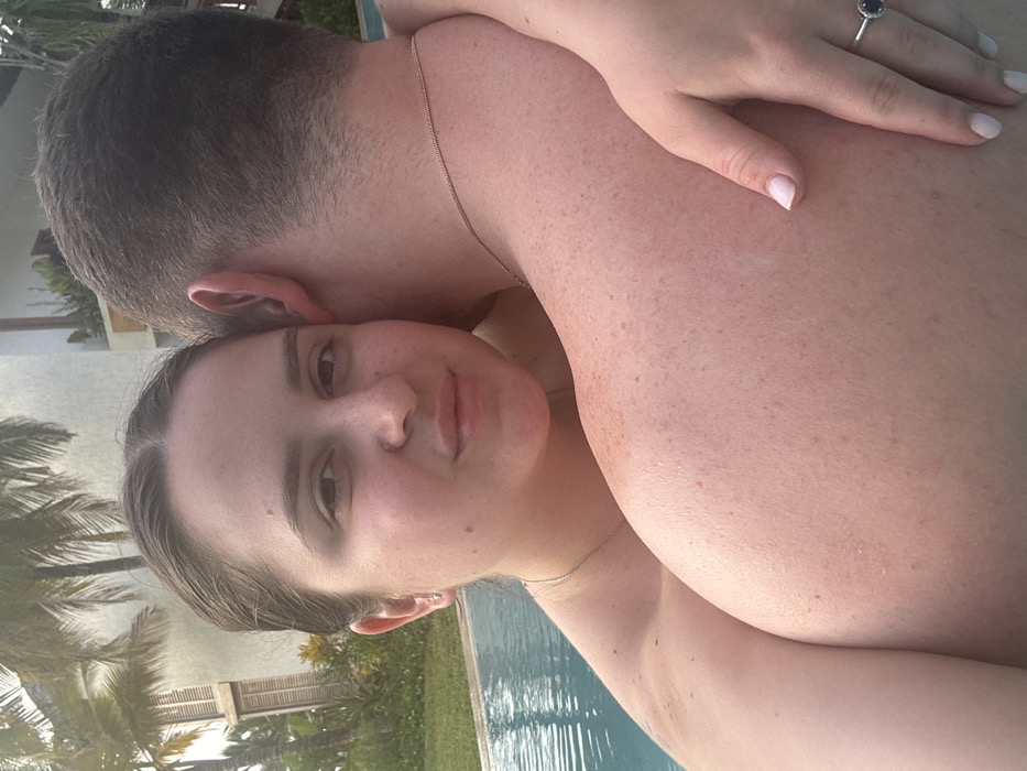 A couple embraces closely. A woman with fair skin, light brown hair, and freckles gazes at the camera. A hand with a silver ring rests on the back of a man with short, dark hair. They appear to be standing at the edge of a pool. Palm trees and the corner of a building are visible in the background.