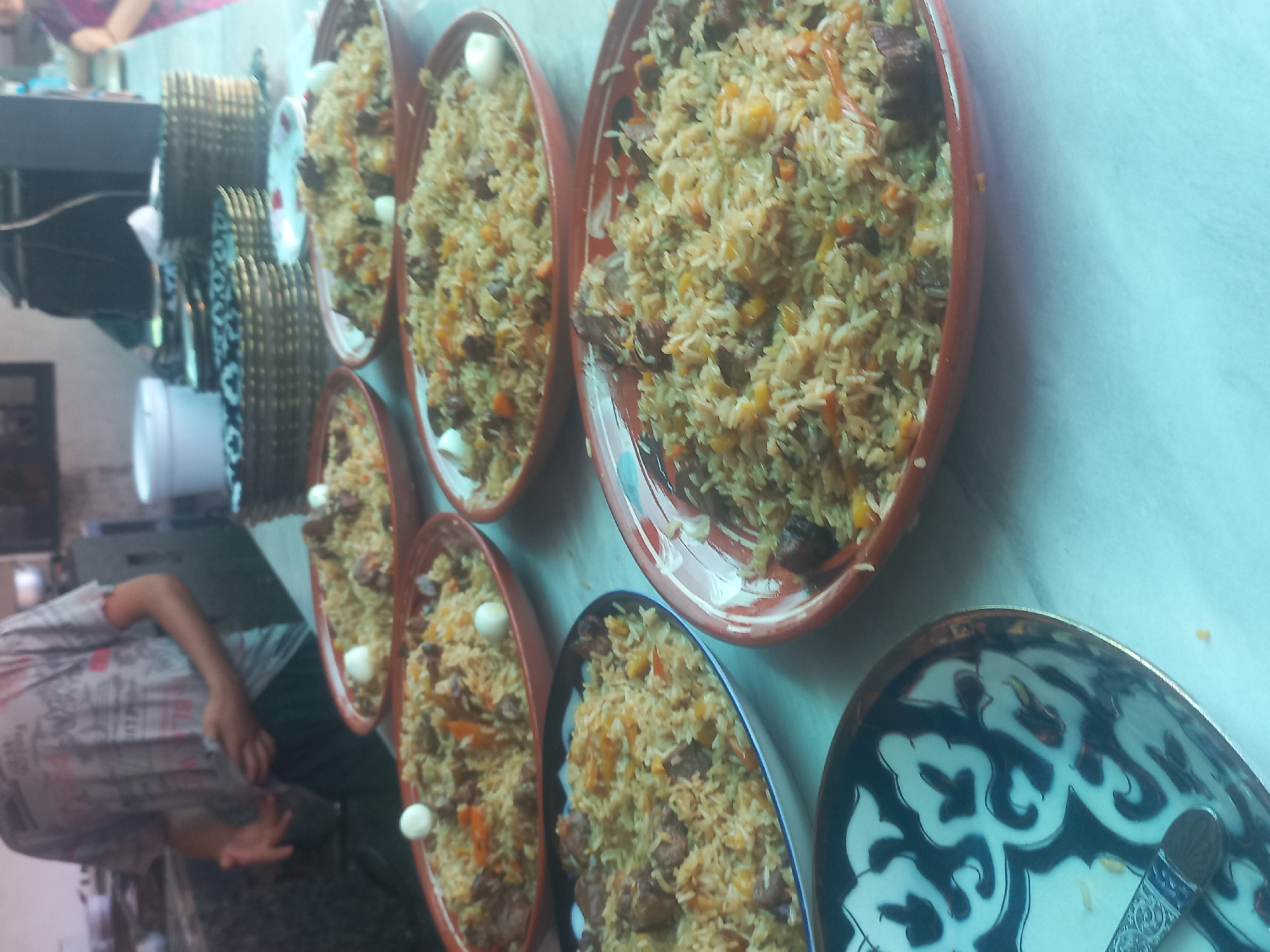 This is a medium-shot, eye-level photograph taken at eye-level inside what appears to be an outdoor restaurant or food stall. The scene is set in Yorkatay, Uzbekistan. A person, partially visible from the waist down, is standing in the background wearing a grey t-shirt and dark pants. The foreground is dominated by several oval-shaped serving dishes filled with pilaf, a traditional Uzbek rice dish. The pilaf contains rice, meat chunks, and shredded carrots, and some dishes are garnished with quail eggs. To the left, a patterned bowl with a blue and white design is visible, containing a fork. In the background, stacked plates and serving baskets are arranged on a table. The overall impression is one of a prepared meal ready to be served.