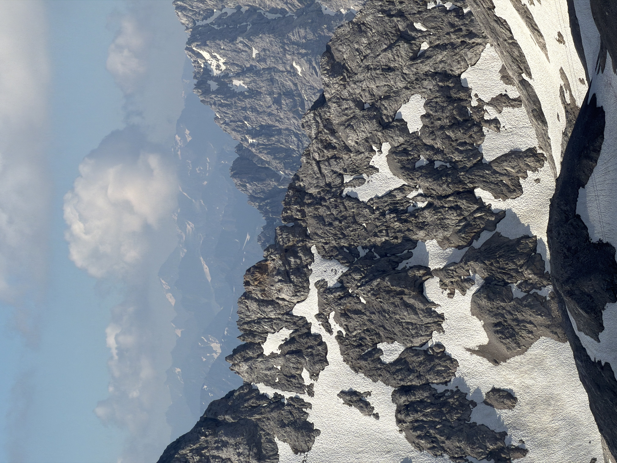 This is a high-angle, portrait-oriented photograph showcasing a rugged mountain landscape. The scene is dominated by steep, rocky peaks partially covered in snow, suggesting a high-altitude environment. The foreground features a close-up view of jagged rock formations interspersed with patches of white snow. These rocks are dark grey and appear weathered, with sharp edges and deep crevices. The snow is not a continuous blanket but rather lies in irregular drifts and patches, highlighting the textures of the underlying rock.
In the background, multiple layers of mountains recede into the distance, their outlines softened by atmospheric haze and a light blue sky. Some of these distant peaks also show touches of snow. The sky on the left side of the image is a clear, pale blue with scattered, fluffy white clouds, indicating fair weather. The overall impression is one of a vast, untamed natural environment.
The lighting suggests it is daytime, with the sun casting shadows on the mountains, emphasizing their three-dimensional form. There are no visible people or man-made objects, reinforcing the wild and remote nature of the location. The image conveys a sense of grandeur and the imposing scale of the mountains.
The location is identified as Garmisch-Partenkirchen, Germany, a region known for its alpine scenery. This type of view is commonly experienced from a high vantage point, perhaps a mountain peak accessible by cable car or hiking, or even from an aircraft. The image does not contain any visible text.