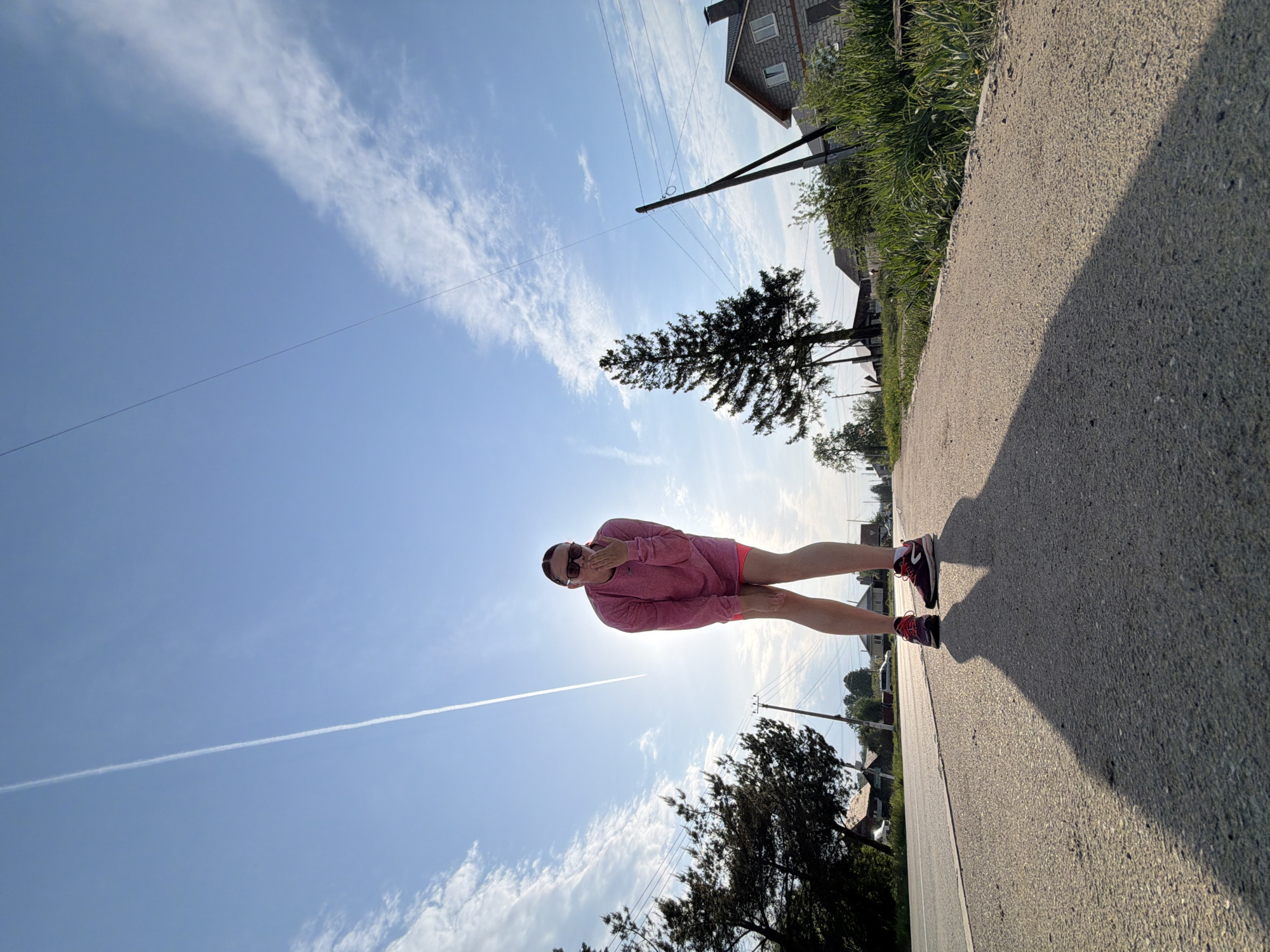 A low-angle, outdoor shot shows a person standing on an asphalt road in Chaikovsky, Russia, with the sky as the backdrop. The person, seemingly a woman, wears a long-sleeved pink shirt, pink shorts, and red and black athletic shoes. She is wearing sunglasses and has her hands raised to her face.
Behind the woman is a blue sky with scattered white clouds and a jet contrail. In the background, there are trees, a house with a gray roof, and utility poles. The asphalt road is textured, with a strip of grass and vegetation to the right of the frame. The angle of the shot creates a perspective where the person appears larger against the sky, with the background elements compressed in the distance.