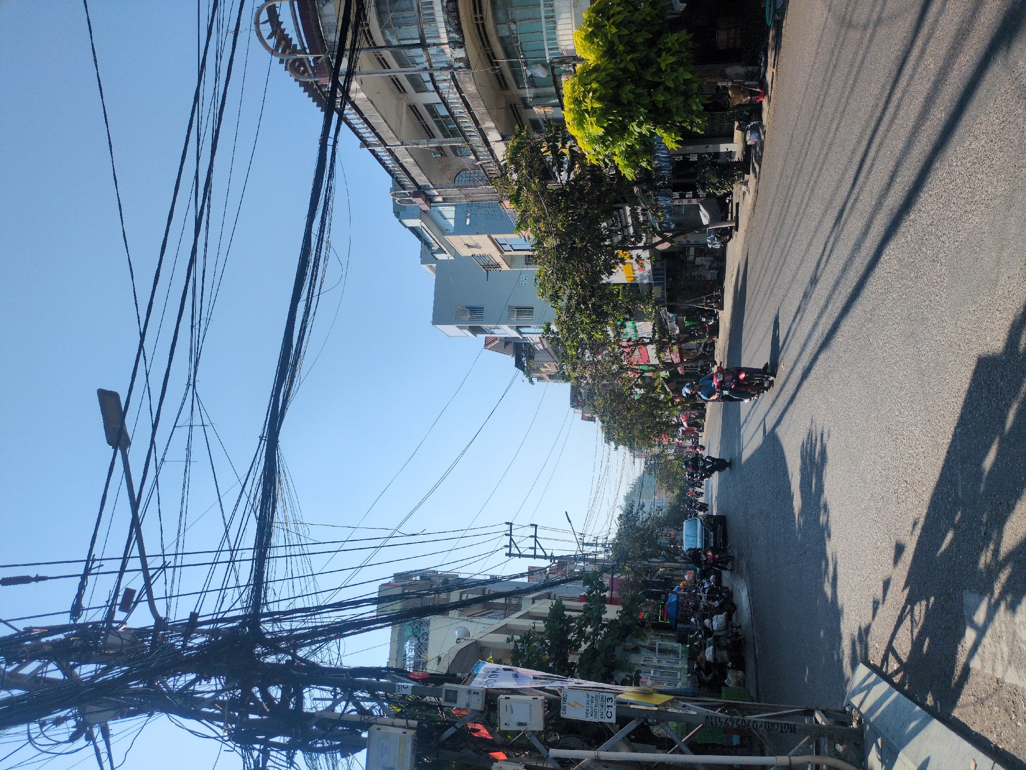 This photograph depicts a street scene in what appears to be a densely populated area, likely in South Quy Nhon Ward, Vietnam. The image is oriented vertically, showcasing a narrow street lined with buildings on either side. A multitude of tangled power lines crisscross the sky above the street.
The street itself is paved and visible vehicles are mainly motorcycles and other similar modes of transportation. The sidewalk areas are crowded with parked motorbikes, suggesting high traffic and a compact urban environment.
The architecture visible consists of multi-story buildings with a mix of residential and commercial facades. Some buildings display signs in Vietnamese. Street vegetation including trees provides some shade to the area.
A closer inspection reveals a sign indicating “C13.” The photo indicates “Electric danger” or high voltage.
