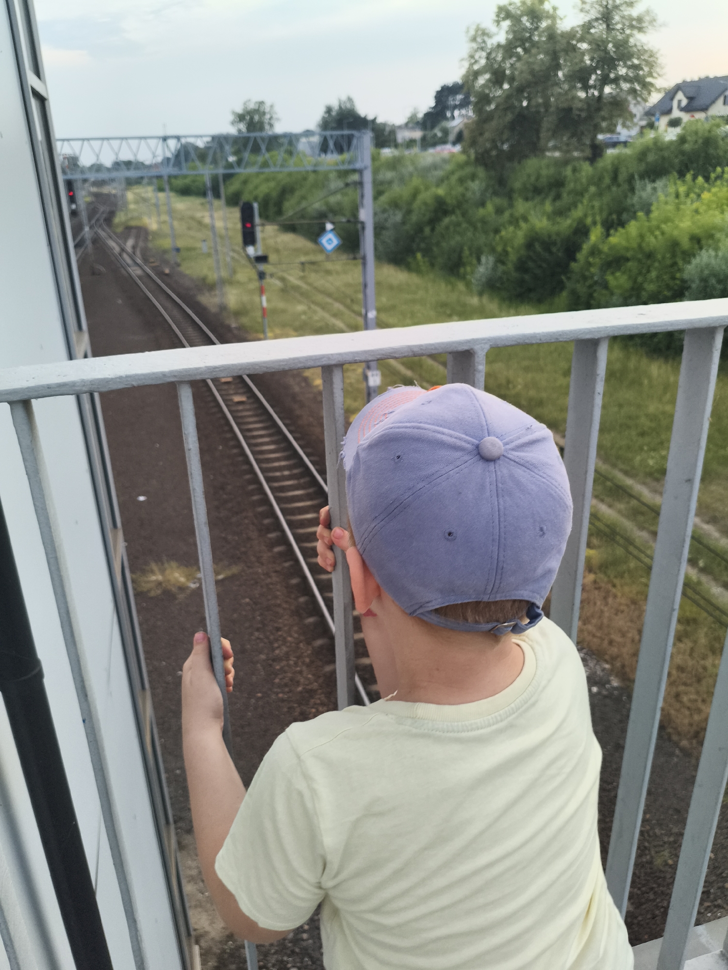 In this medium shot, a young person wearing a light yellow t-shirt and a purple baseball cap is standing at a railing, looking out at a railway line. The viewpoint is from behind the person, focusing on their back and the cap. The railway tracks stretch into the distance, curving to the left. A railway signal with a red light is visible on a pole to the right of the tracks, and a blue diamond-shaped sign is positioned behind it.
Overhead, a metal structure supporting electrical wires for the trains spans across the tracks. The surrounding area is lush with green trees and vegetation, with a few houses visible in the background on the right. The sky is overcast, suggesting it might be late afternoon or early evening, and the weather appears to be mild. The person's hands are gripping the metal railing, and their posture suggests they are observing something intently on the railway. The scene evokes a sense of quiet observation and perhaps anticipation, as if waiting for a train. The location context places this scene in Nowy Dwór Mazowiecki, Poland.