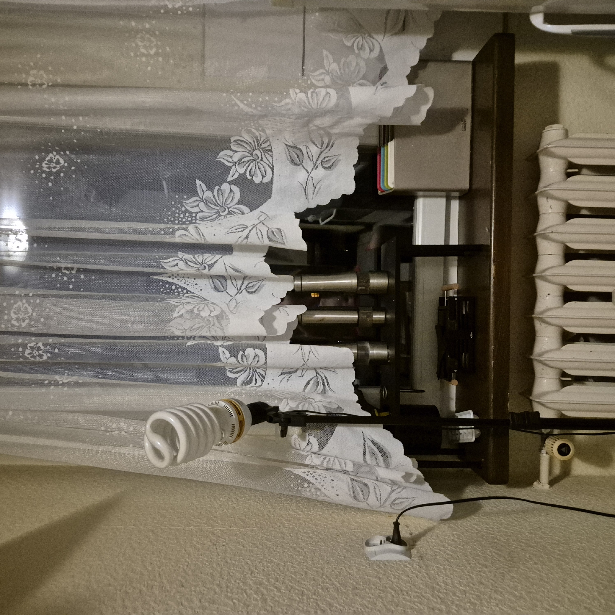 A floor lamp with a spiral fluorescent bulb is set up next to a window with sheer white curtains. The lamp is on a black tripod stand and is plugged into a white electrical outlet on the textured wall. Below the window is a dark shelf holding a few items, including what appear to be salt and pepper grinders, and a box containing colored items. A white radiator is visible below the shelf. The scene appears to be indoors, possibly a home or office, in Kępno, Poland.