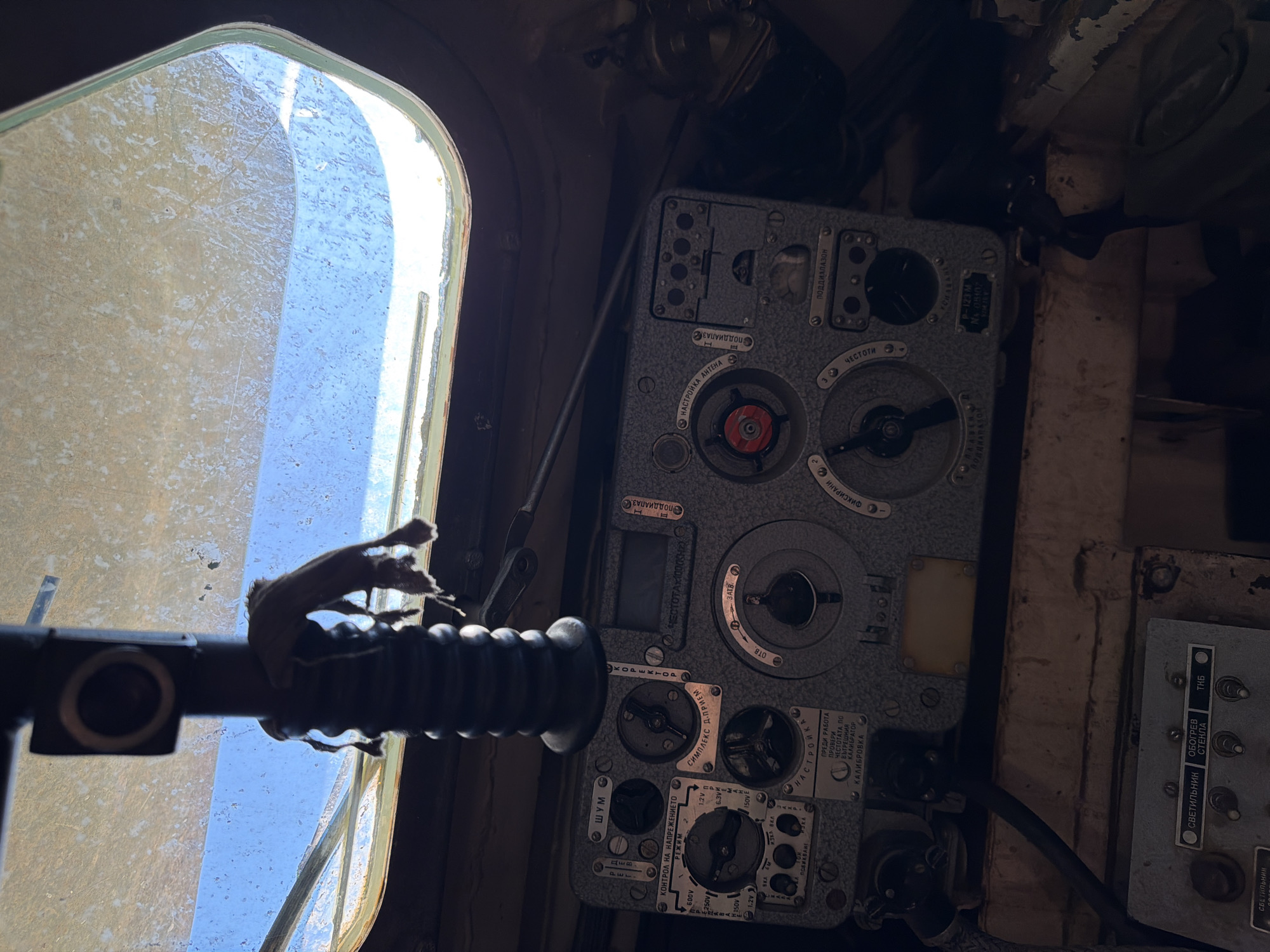 The image shows a close-up view of the interior of a vehicle, likely a military vehicle, with a focus on a control panel. The panel is gray and covered with various knobs, dials, and labels in Cyrillic script. The labels suggest functions related to radio communication, voltage control, and tuning. The overall environment appears to be somewhat aged and dusty. In the background, a window is visible, showing a bright sky, possibly indicating daylight. A dark, ribbed mechanism is positioned above the control panel, and a small, tattered piece of fabric is attached to it. The overall impression is one of outdated technology and a functional, albeit utilitarian, interior. The location in Beliakovets, Bulgaria, is not visually evident from the image itself.