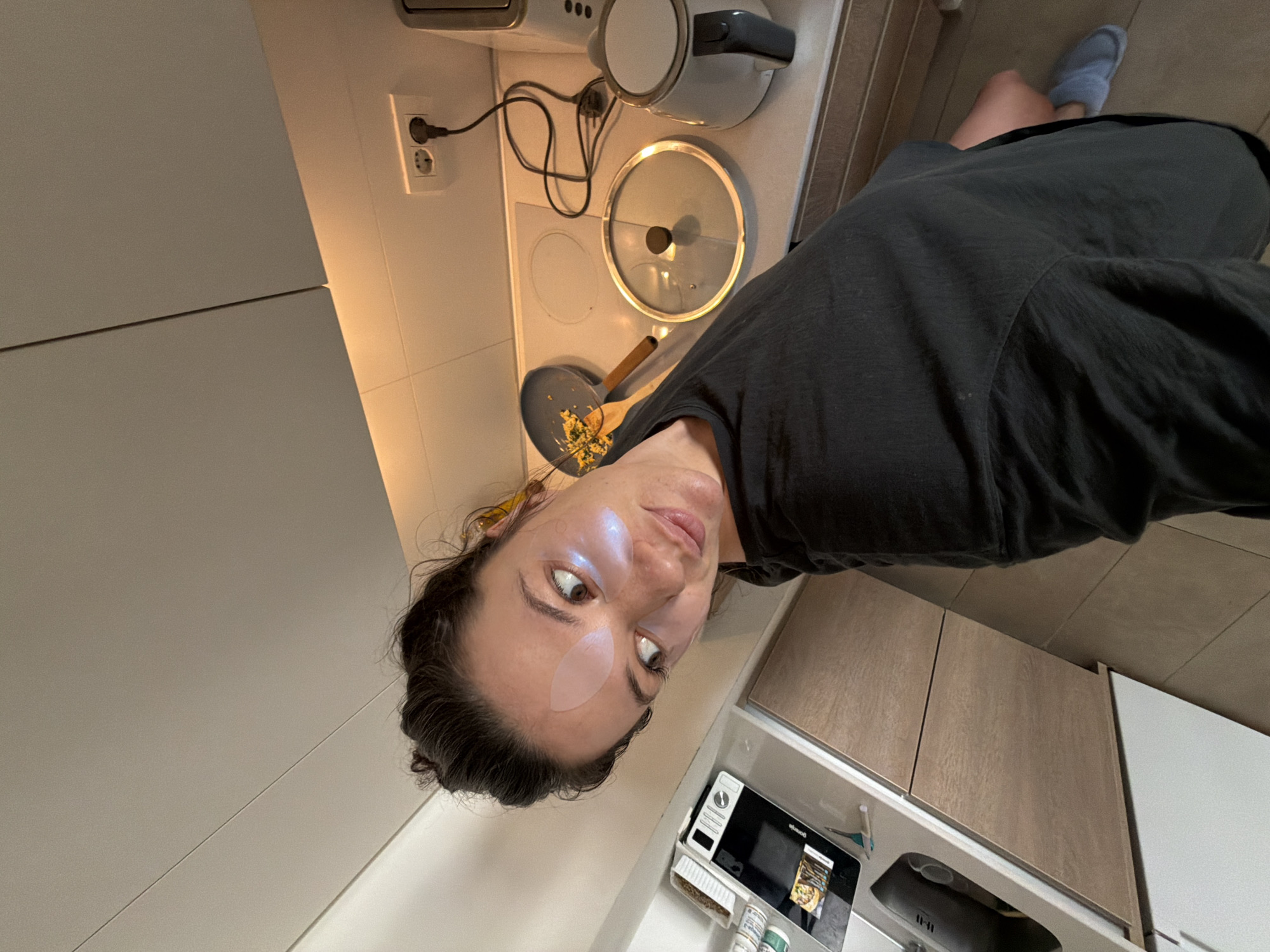 A woman is in a kitchen in Belgrade, Serbia. She has a light pink patch on her forehead. She is wearing a dark t-shirt. On the stovetop behind her is a kettle, a pan with food cooking, and a pot lid. The kitchen has white and light brown cabinets, a microwave, and a sink. The woman is looking directly at the camera.