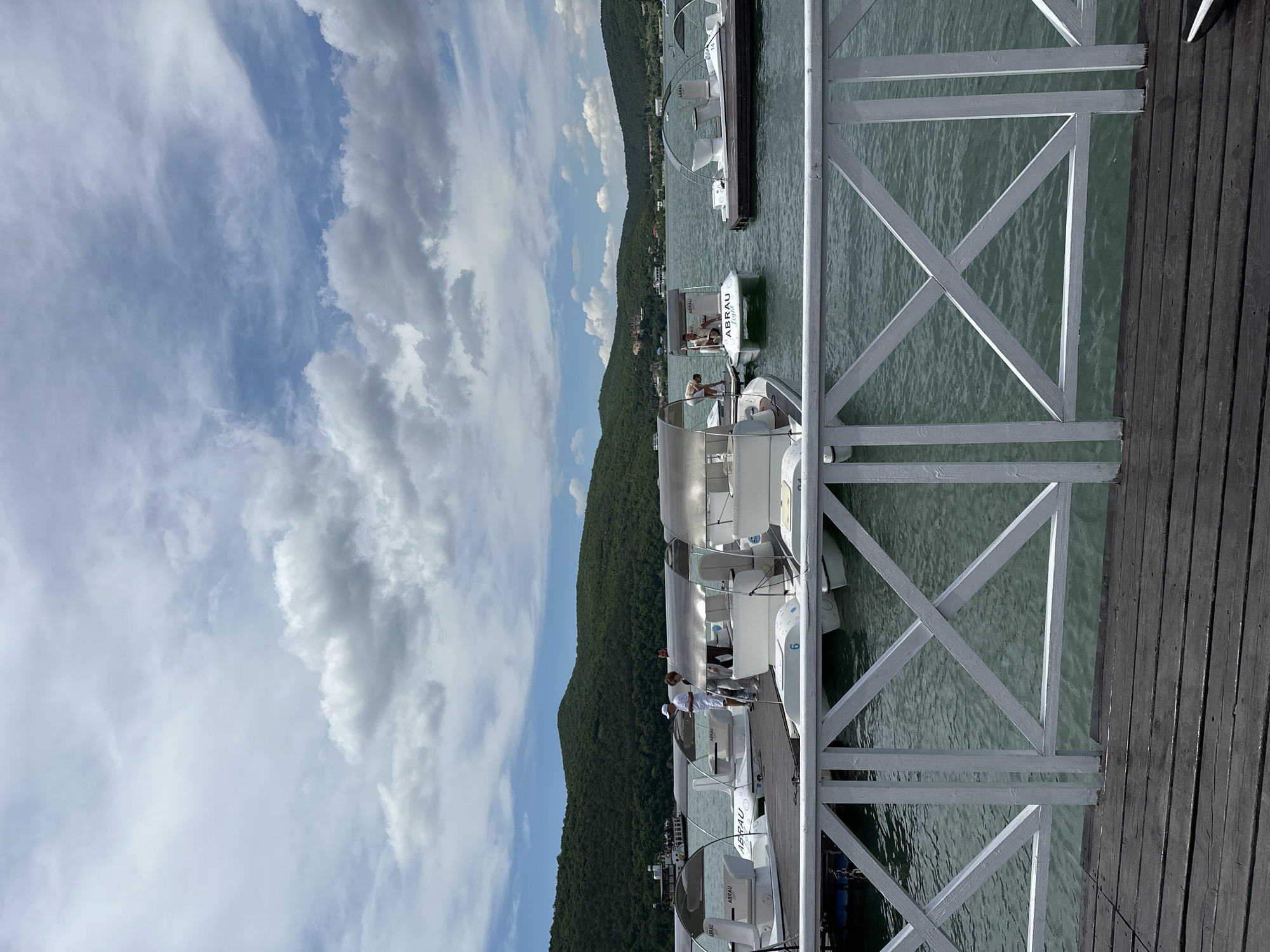 A vertical, eye-level outdoor shot captures a serene waterside scene in Abrau-Dyurso, Russia, on a partly cloudy day. In the foreground, a weathered wooden pier stretches out, bordered by a white, cross-latticed railing. Beyond the railing, a calm, greenish body of water reflects the sky, with several small, white pedal boats moored to a wooden dock.
On the dock, a few individuals are visible. One person, wearing a white t-shirt and cap, is seen tending to a pedal boat on the left. Another individual, also in a light-colored shirt, appears to be standing near the center, talking with someone else. A few other figures are further down the dock, possibly preparing for or disembarking from the boats. The boats themselves are modern and compact, each with a canopy for shade. One boat is marked with the number "6."
The middle ground of the image is dominated by the lake and the floating dock. In the background, a lush, green hillside rises steeply, covered in dense trees. At the very top of the hill, a cluster of buildings, possibly a resort or village, can be seen. The sky above is a mix of pale blue and white clouds, some of which are quite puffy and dramatic. The overall impression is one of a tranquil recreational area by the water.